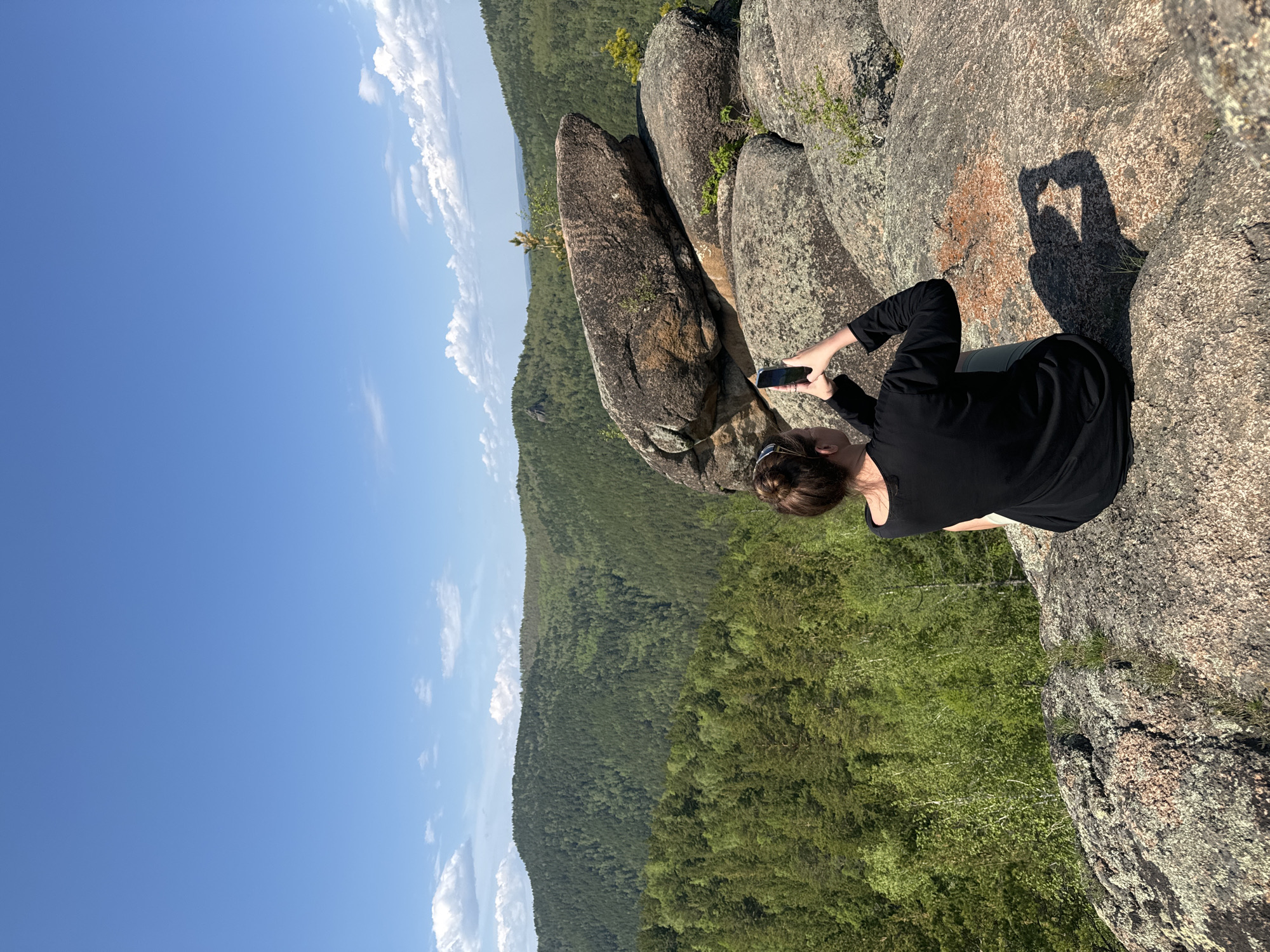 The image depicts a woman on a rocky precipice, possibly taking a photograph with a smartphone. She is positioned on a rock formation overlooking a forested valley and a clear blue sky with scattered clouds. The location is likely in or near Krasnoyarsk, Russia, based on the geographical features and rock formations characteristic of the Stolby Nature Reserve. The woman wears a black long-sleeved shirt and her hair is pulled back in a bun. The rock surface where she is seated is covered in lichen and small plants. A shadow in the shape of a heart is cast on the rock face next to her.