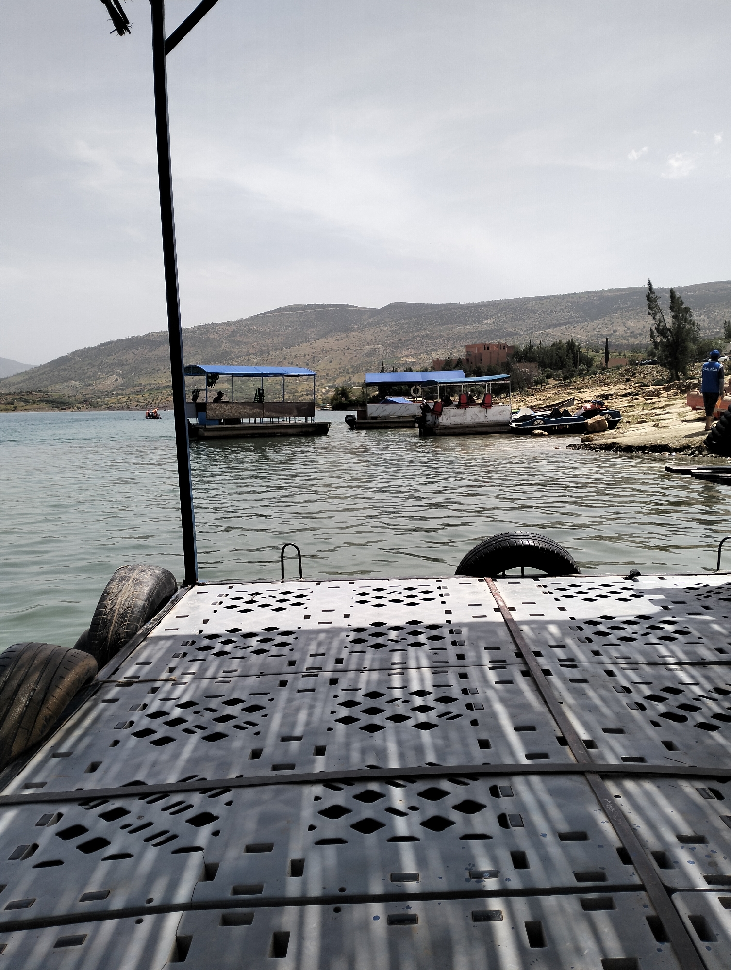 The image depicts a tranquil scene at Bin El Ouidane, Morocco, featuring a lake with several boats. A floating platform constructed of gray plastic modules secured with metal bands is in the foreground, with tires attached to the side acting as bumpers. In the water, two covered pontoon boats are visible, with several people visible on one of the boats. A small motorboat is docked near the shoreline, where people are also gathered. The background reveals a hilly landscape with sparse vegetation and buildings. The sky is overcast, suggesting a muted, diffused light. The overall impression is a tourist or recreational area on a relatively calm day.