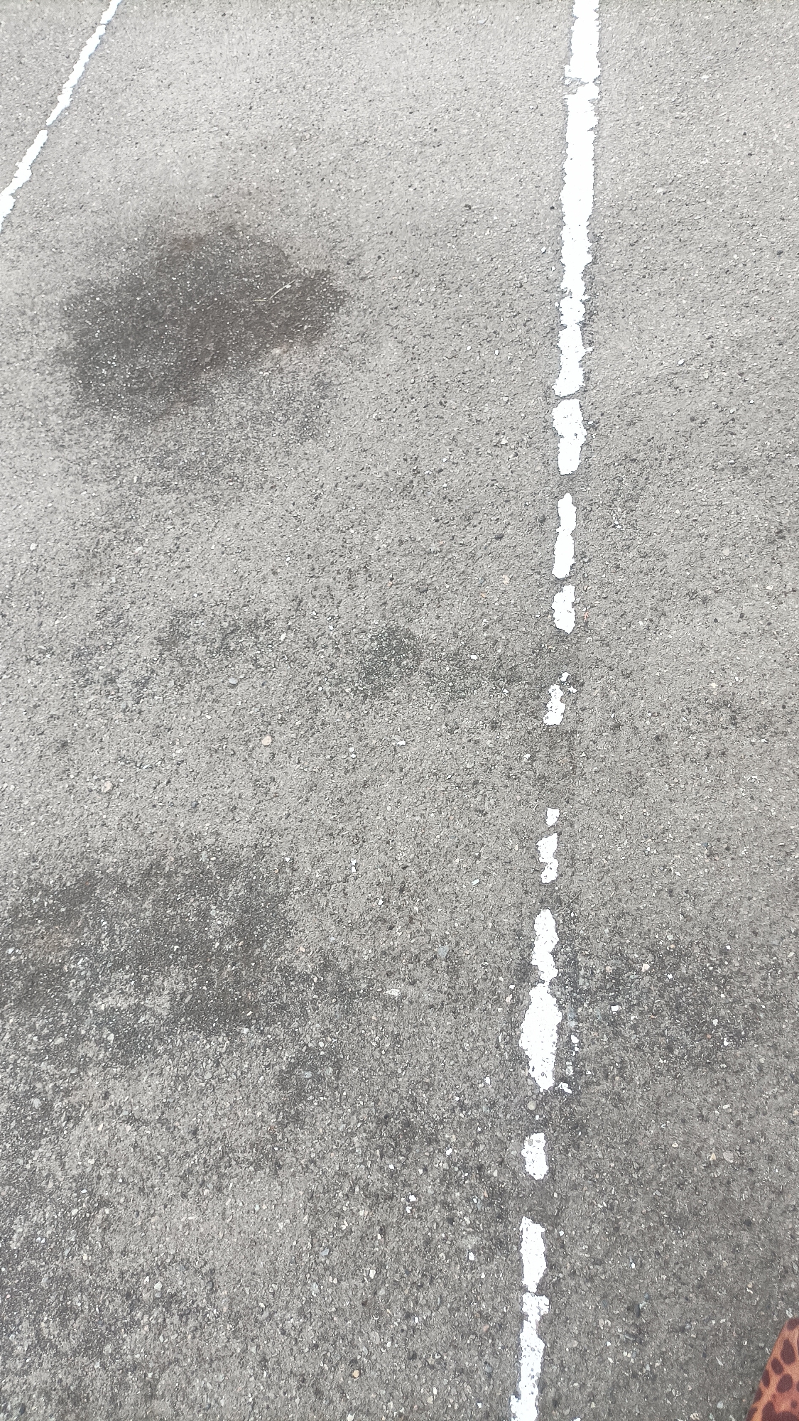 The image shows a close-up, high-angle view of an asphalt surface, likely a roadway or parking lot. A broken white line, common for lane division or parking stall demarcation, runs vertically down the right side of the frame, with segments of white paint chipped and uneven. To the left of this line, a dark, irregular stain, possibly oil or water, is present on the asphalt. The asphalt itself appears weathered and textured, with visible aggregate. In the bottom right corner, a small portion of a patterned fabric, possibly from clothing or a bag, is visible. The scene is devoid of any discernable landmarks that would pinpoint its location within Vanadzor, Armenia, or any other city. The environment is generic and consistent with urban or suburban outdoor paved areas. There are no people or discernible actions taking place.