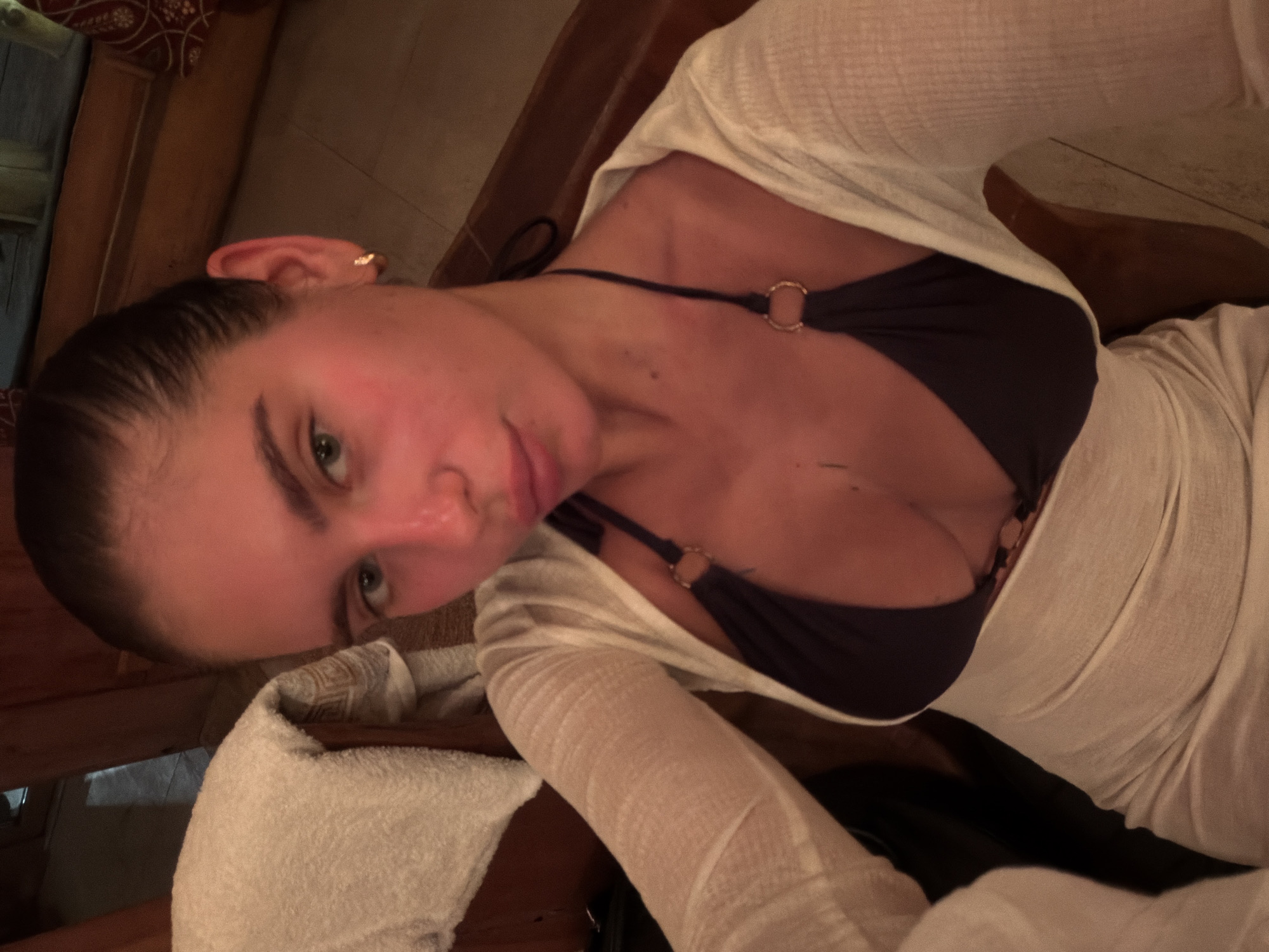 The image captures a young woman lying in what appears to be a warm, dimly lit indoor setting, possibly a sauna or spa, characterized by prominent wooden paneling in the background. Her hair is pulled back, and she is looking directly at the camera with a calm, slightly pouting expression.
She is wearing a dark-colored bikini top, visible beneath a sheer, light-colored, long-sleeved cover-up or top. The bikini top features gold-toned ring details on its straps. A small golden hoop earring is visible on her left ear. In the lower left foreground, a white towel is draped over a wooden surface.
The lighting suggests an evening or nighttime setting. The overall mood is relaxed and intimate, indicative of a leisure or self-care activity. The photograph appears to be a close-up selfie due to the direct gaze and perspective. No visible text is present in the image.