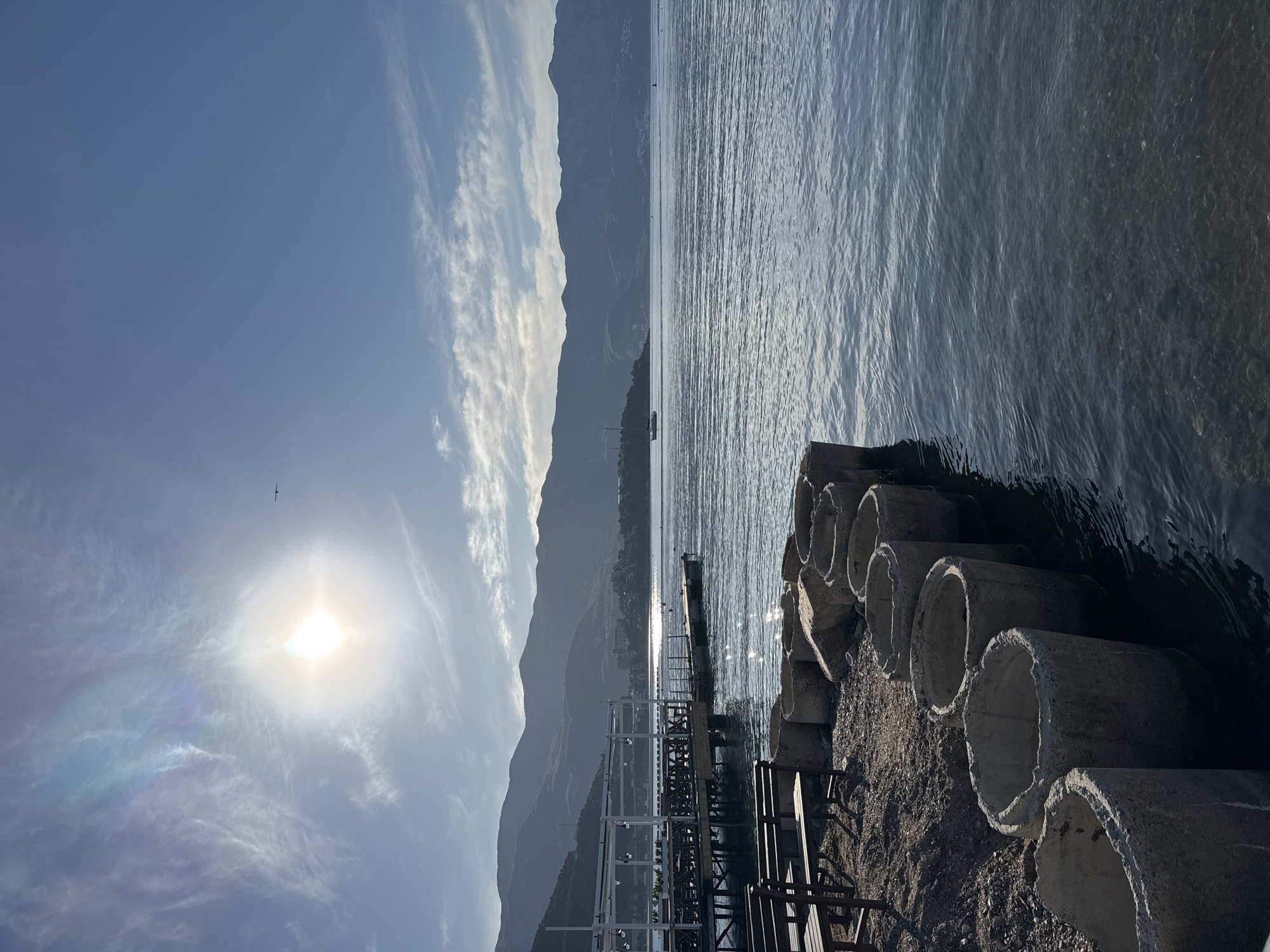 The image depicts a coastal scene in Budva, Montenegro. In the foreground, there are several stacked concrete pipes on a gravelly area near the water's edge. A metal pier extends into the water, with some sections partially submerged. In the middle ground, there is a body of water, possibly the Adriatic Sea, reflecting sunlight. A boat is visible in the distance. The background features mountainous terrain and a partly cloudy sky with a bright sun, which includes a visible bird flying in the sky.