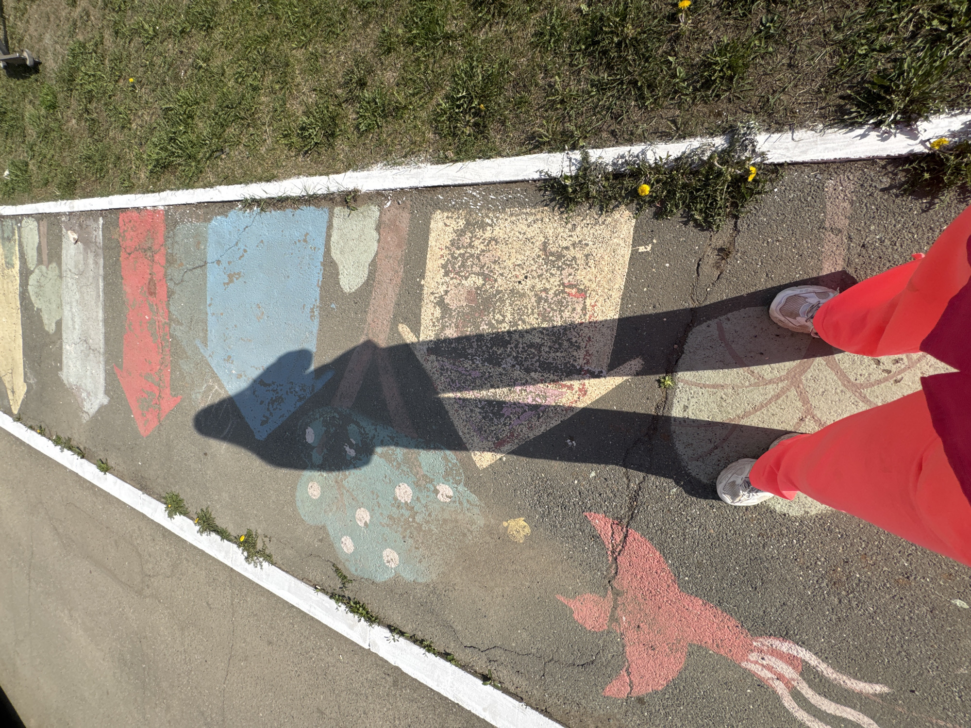 A photograph taken outdoors shows a section of cracked asphalt decorated with colorful chalk drawings of arrows, geometric shapes, and a bird. A person wearing bright pink pants and white sneakers stands on the painted asphalt, casting a dark shadow. The location is likely Zav'yalovo, Russia. Adjacent to the painted asphalt is a strip of grass and a concrete sidewalk.