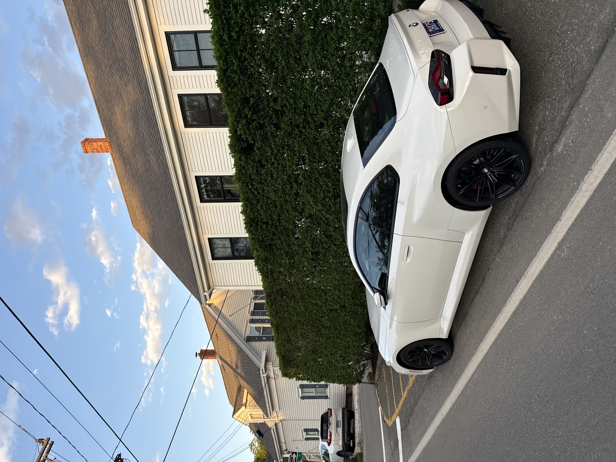 The image depicts a white BMW parked on a street in Provincetown, United States of America. The car is positioned next to a tall, dense green hedge that runs alongside a white, two-story house with black-framed windows. The house has a gray roof with a red brick chimney. Another house is visible behind the first one. Power lines stretch across the upper left portion of the frame, against a backdrop of a blue sky dotted with light clouds. The car is parked on a paved street marked with white and yellow lines.