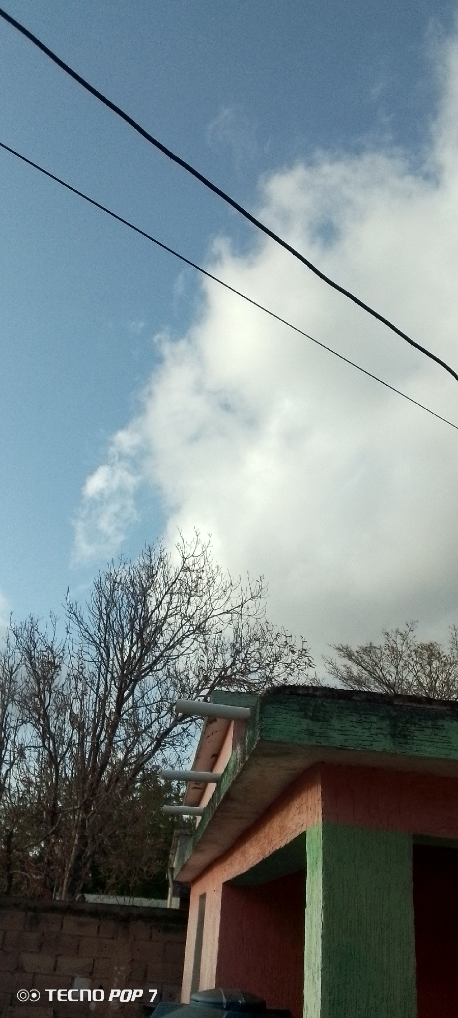 The image shows a partly cloudy sky over a small building in Altagracia, Venezuela. Two black electrical lines cross the upper portion of the frame. In the middle ground, there are leafless trees. The building in the foreground has a green and orange facade with white pipes extending from the roof. The sky is visible through the building’s openings. A brick wall is partially visible on the left side of the frame. A "TECNO POP 7" watermark is in the lower left corner.