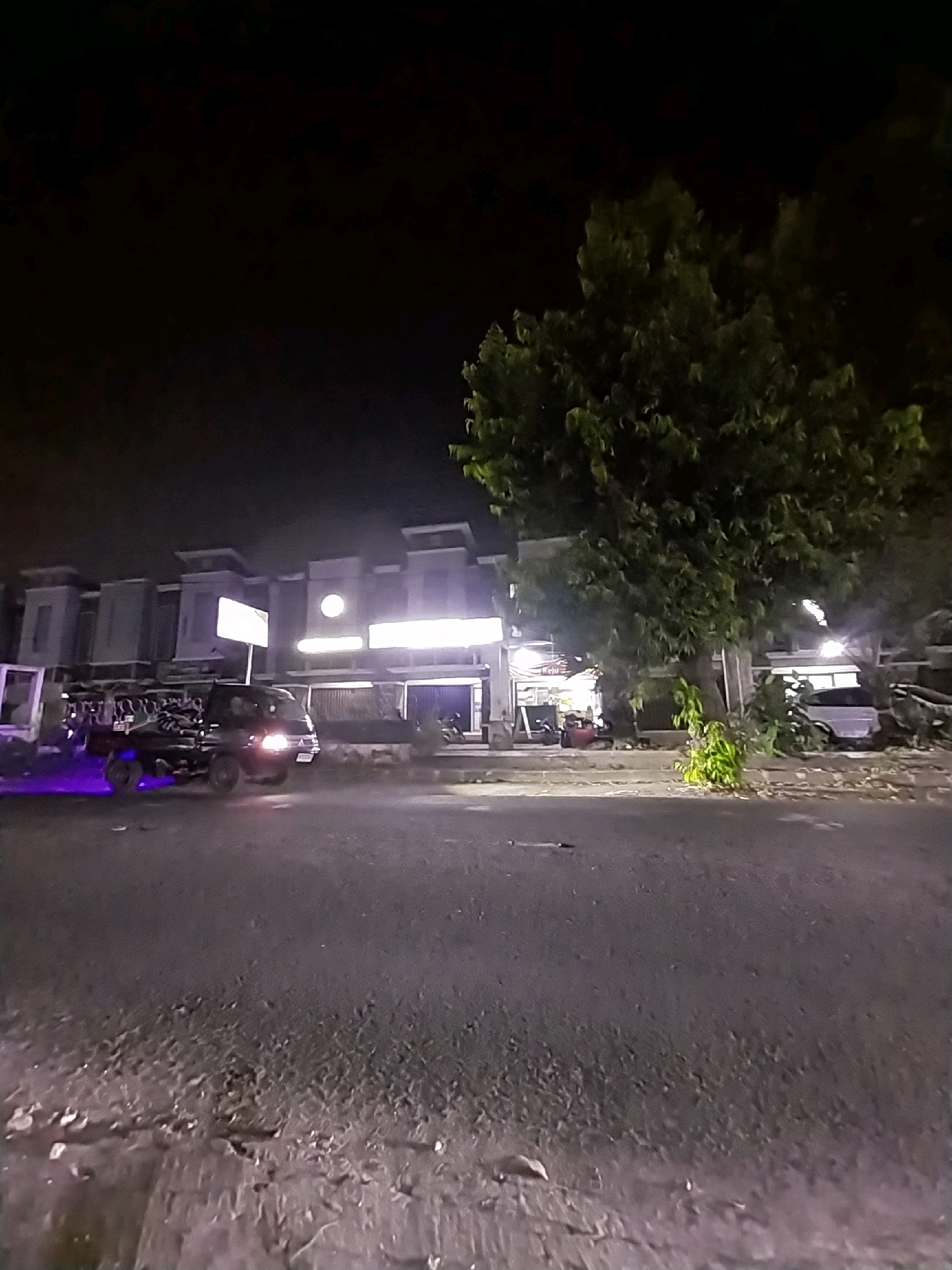 The image depicts a nighttime street scene in Tlogoadi, Indonesia. A dark-colored truck is parked on the left side of the road, illuminated by purple underglow lights. In the background, there are several multi-story buildings with illuminated windows and signage, suggesting commercial establishments. A large tree with dense foliage stands to the right of the buildings. The road surface appears to be asphalt with some debris. The sky is dark, indicating nighttime. There are no people clearly visible in the image.