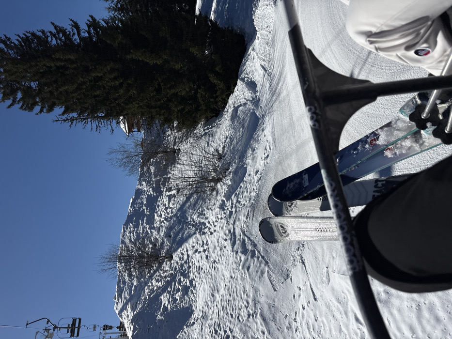 The image depicts a first-person view from a ski chairlift ascending a snow-covered mountain in Manigod, France, under a clear blue sky. In the immediate foreground, the safety bar and footrest of the chairlift are visible, with two pairs of skis dangling beneath. The left pair consists of a silver/white and a blue/black ski, both appearing to be Rossignol brand, attached to dark-colored ski boots worn by a person in dark ski pants. To the right, part of a second person's right leg, wearing light grey ski pants with a small red, white, and blue emblem, and a blue ski with a similar Rossignol design are visible.
Below the chairlift, a snowy slope descends, marked with tracks and shadowed undulations. Sparse, leafless deciduous bushes and small trees are scattered across this slope. To the right, a dense cluster of tall evergreen coniferous trees lines the hillside. Partially obscured by these evergreens and situated higher up on the slope is a wooden chalet-style building with a snow-covered roof. In the upper left quadrant, empty chairlift seats are visible higher up the line. The scene is illuminated by strong sunlight, casting distinct shadows on the snow.