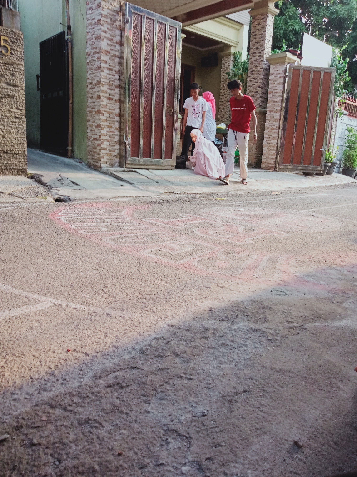 In East Jakarta, Indonesia, in front of a house with a beige facade and brown metal gates, a group of four individuals are gathered on a paved driveway and street. The driveway is marked with chalk drawings, predominantly pink, suggesting a children's game like hopscotch or a drawing.
Two young men stand near the entrance, one wearing a white t-shirt with text and the other a red t-shirt with "POWER PLAYER" printed on it. A young woman, dressed in a pink hijab and a white garment, is seated on the ground, appearing to be interacting with the chalk drawings or something on the ground. Behind her, another person, partially obscured, is also present. The environment includes potted plants and a rough textured wall of the building. The lighting suggests it is daytime, with shadows indicating the sun's position. The street itself is rough and textured.