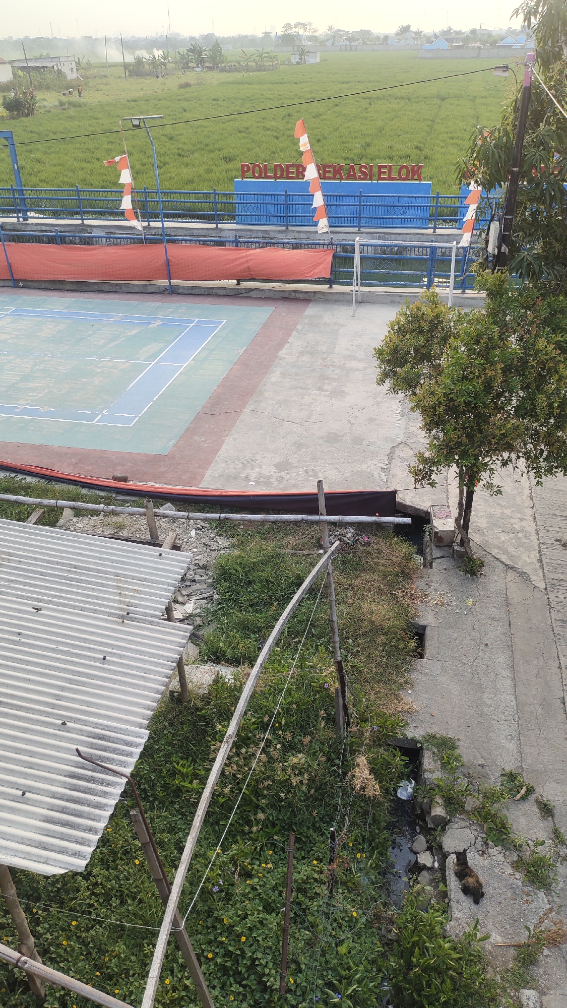 The image depicts an outdoor scene that appears to be in Jalenjaya, Indonesia. In the background, there is a lush green field, likely rice paddies, bordered by a blue fence. A red sign with white text reads "POLDER BEKASI ELOK" and is accompanied by Indonesian flags. In the foreground, a portion of a badminton court is visible, with blue and green playing surfaces marked by white lines. Adjacent to the court is a red net and a small white soccer goal. To the right of the court, a paved pathway runs alongside a ditch filled with water and debris, with a small tree growing nearby. On the left side of the image, a corrugated metal roof of a structure is visible, supported by wooden beams and surrounded by dense green vegetation and some yellow flowers. The overall environment suggests a rural or semi-urban area with recreational facilities and natural elements.