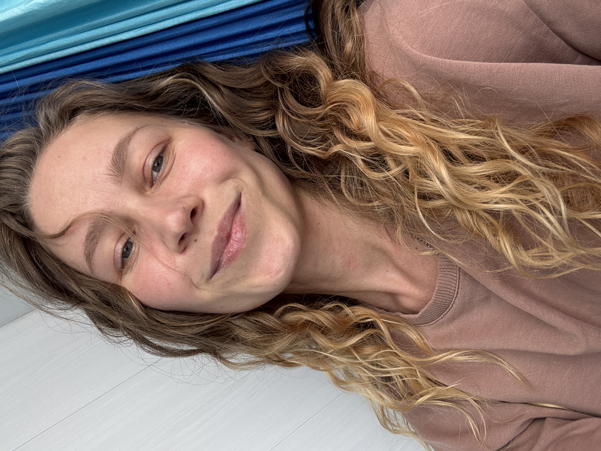 A woman is lying down, angled diagonally in the frame. She has wavy, brown and blonde hair, and is wearing a light brown sweatshirt. Above her is a striped blue and turquoise fabric. Below her is a white, possibly wooden, surface. The location, Kladovitsy, Russia, is not visually discernible from the image.