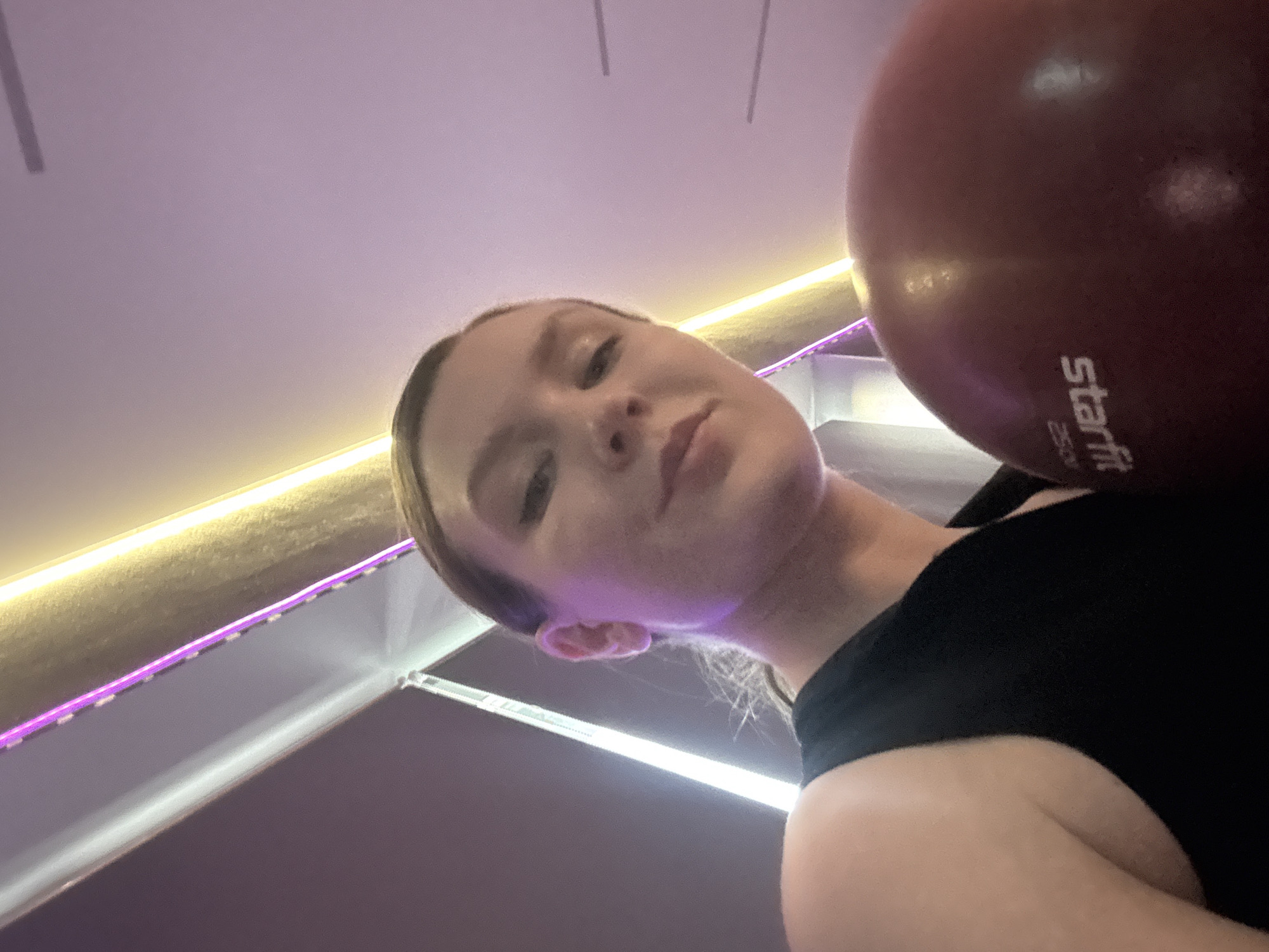 A low-angle selfie shows a woman with fair skin and light brown hair pulled back, holding a red "Starfit" brand exercise ball against her right shoulder. She is wearing a black top. The background features a ceiling with recessed lighting, including a strip of purple LED lights. Based on the "Starfit" branding on the ball, this may be a fitness or sports facility. Without further information, it's impossible to confirm the precise location in Izhevsk, Russia.