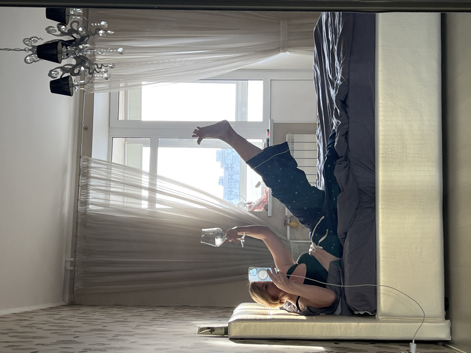 The scene depicts an interior apartment view in Saint Petersburg, Russia, captured via a mirror reflection. An adult female with light-colored hair is lying supine on a bed, oriented towards the left. She wears a dark green sports bra and dark blue pajama bottoms with a white star pattern. Her left hand holds a white smartphone with a clear case, connected by a grey charging cable, pointed towards her face. Her right hand is raised, holding a stemmed glass containing a clear liquid. Her right leg is bent with the foot resting on the bed, while her left leg is extended vertically with the foot flexed.
The bed consists of a cream-colored upholstered headboard and frame, dressed with dark grey linens. The room's left wall features light-toned, patterned wallpaper. A prominent white-framed window, partially obscured by sheer white curtains, reveals a bright, overcast sky over a distant cityscape of multi-story residential buildings, consistent with urban Saint Petersburg. A white radiator is visible below the window sill, which also holds a small decorative item. A chandelier with black shades and clear crystal pendants hangs from the white ceiling.