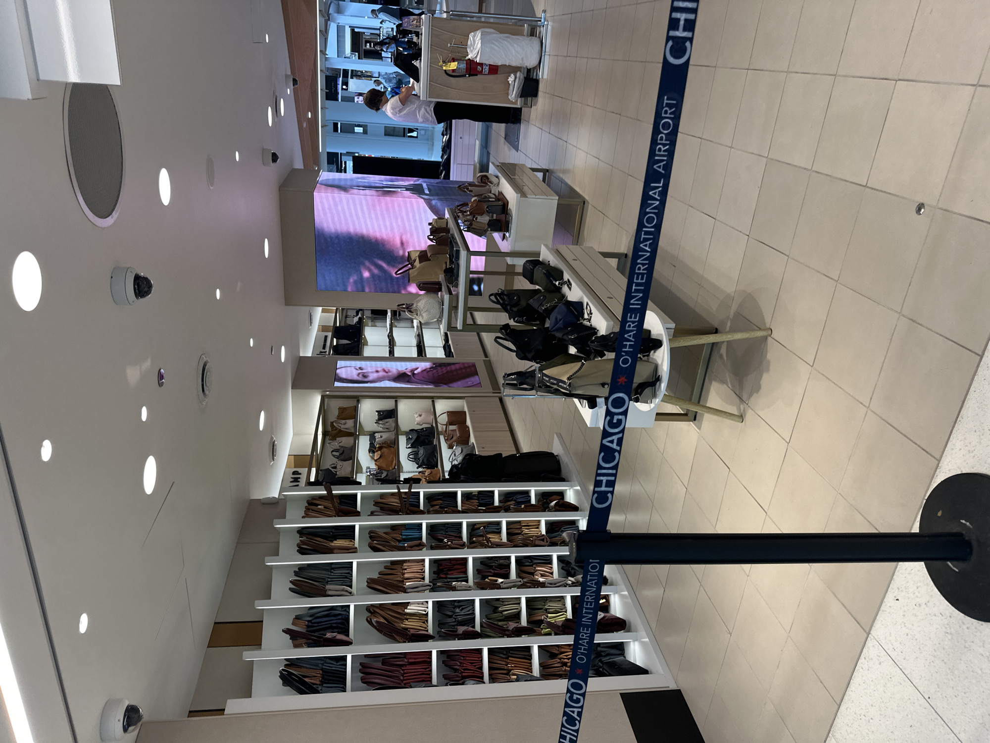 The image shows the interior of a retail store within O'Hare International Airport in Chicago, Illinois, United States of America. The store specializes in handbags and accessories, with numerous bags displayed on white shelving units. A blue stanchion with a tape barrier that reads "CHICAGO O'HARE INTERNATIONAL AIRPORT" runs across the foreground, indicating a queue or entrance. In the background, a person, likely an employee, is standing behind a counter, interacting with a customer. A large digital screen displays an advertisement featuring a woman's face. The store has a modern aesthetic with tiled flooring and recessed lighting.