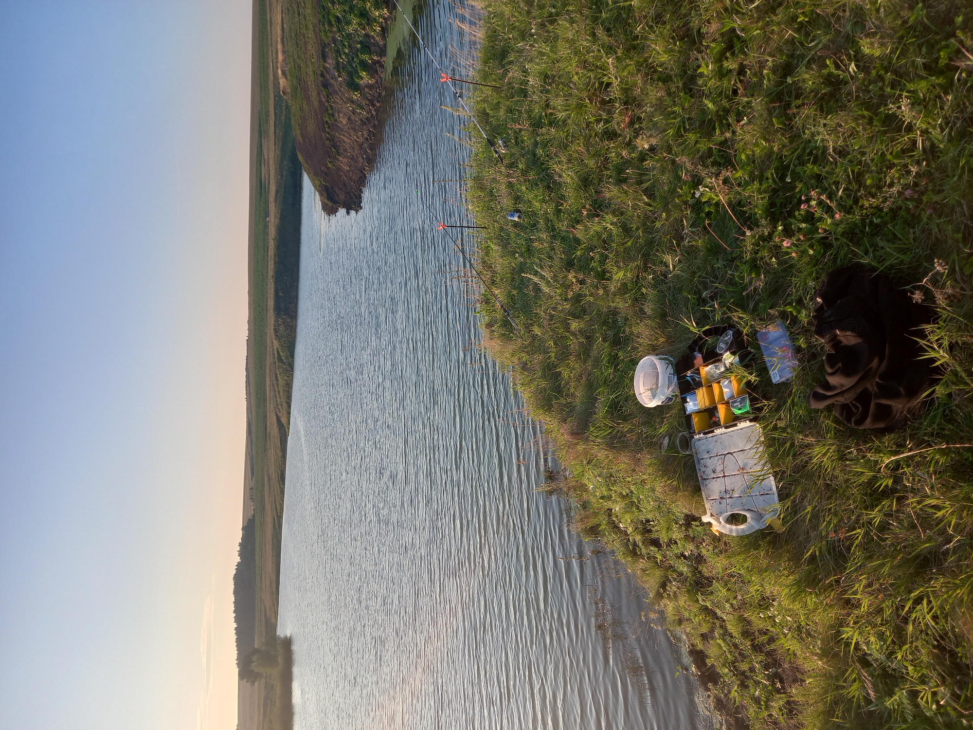 The image shows a tranquil natural scene on the outskirts of Polevye Ineli, Russia, beside a calm body of water. The sun is low in the sky, casting a warm, golden light across the landscape, creating long shadows and highlighting the texture of the grass and water. On the grassy bank of the water, fishing equipment is laid out, including two fishing rods propped up by orange holders, a tackle box filled with various fishing lures and accessories, and a white bucket. A dark fabric, possibly a jacket or blanket, is resting on the grass near the tackle box. The water is a deep blue, with gentle ripples on its surface, reflecting the clear, pale blue sky. The opposite bank of the water is lined with green vegetation and more distant fields, suggesting a rural or semi-rural setting. The overall impression is one of peaceful solitude, likely a scene of a fishing trip or a moment of quiet contemplation in nature.