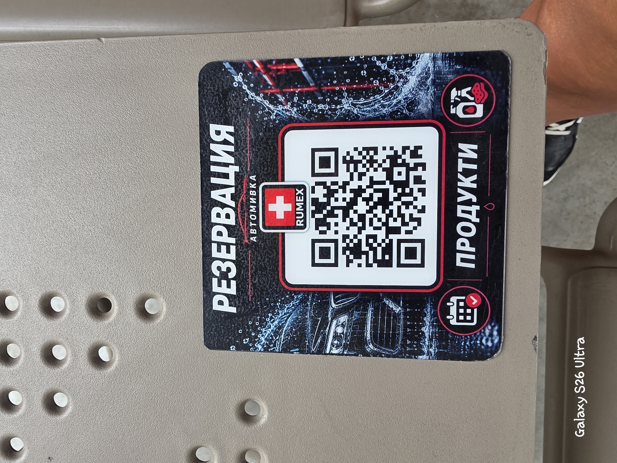 The image shows a sticker on a beige plastic surface, possibly part of a car wash facility. The sticker features a QR code, a red cross logo with "RUMEX" text, and text in Cyrillic: "РЕЗЕРВАЦИЯ" (Reservation) and "ПРОДУКТИ" (Products). There are also icons depicting a calendar with a checkmark and car cleaning supplies. The background of the sticker shows a stylized image of a car with water splashes. The bottom of the image has a watermark reading "Galaxy S26 Ultra". The environment suggests an outdoor or semi-outdoor setting, potentially a public space in Plovdiv, Bulgaria, given the Cyrillic text, although no specific landmarks are visible. A person's leg and foot wearing a black sneaker with white stripes are partially visible in the lower right corner of the image.