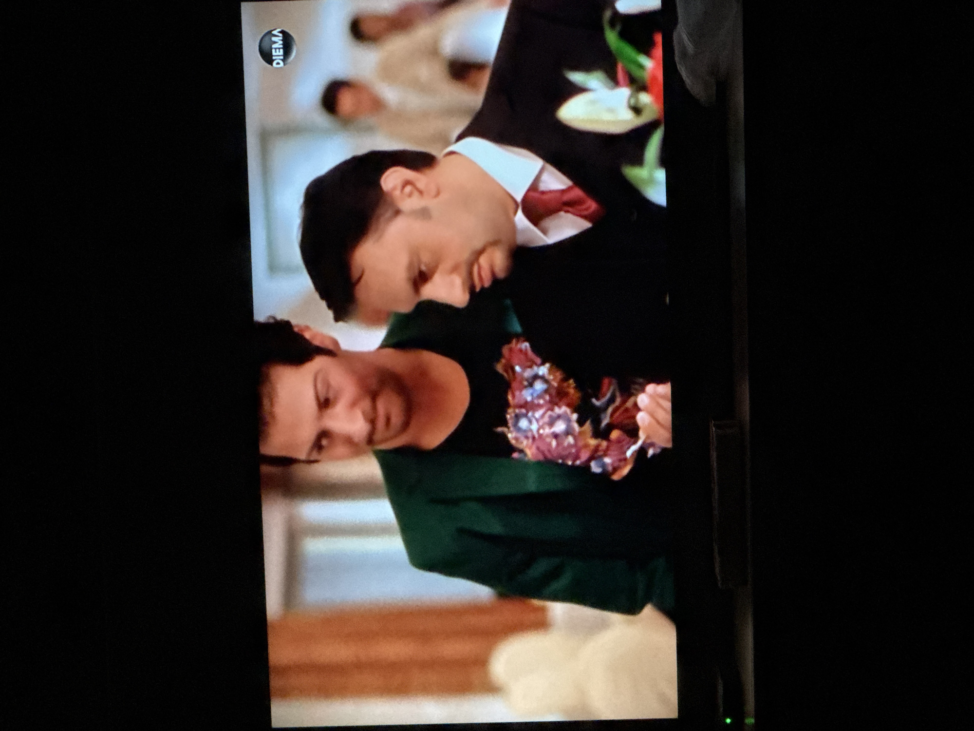 Here is a description of the image:
The image is a shot of a television screen displaying a scene with two men. One man is wearing a suit with a white shirt and red tie, while the other is wearing a green jacket over a black shirt with a floral design. The man in the suit is leaning towards the man in the green jacket, possibly embracing or comforting him. In the background, there are other people in formal attire, suggesting a wedding or formal event. The setting appears to be indoors. A "DIEMA" logo is visible in the upper left corner of the screen. There is no location information in the image.
