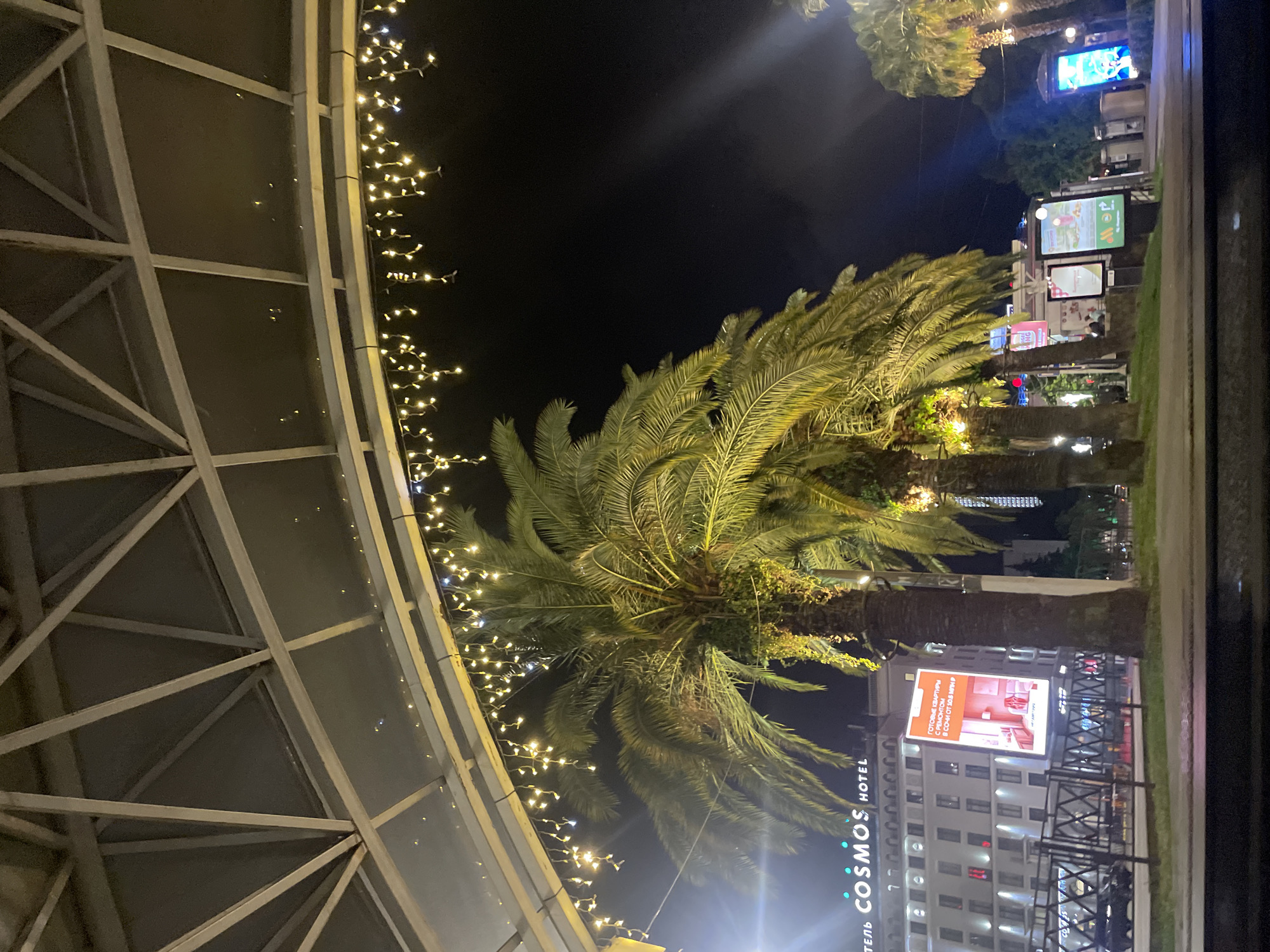 This is a nighttime exterior shot in Sochi, Russia, featuring several palm trees in the foreground. Behind the palm trees, a large hotel with the sign "COSMOS HOTEL" is visible. A billboard on the hotel displays an advertisement for apartments with renovations. String lights are hung from an overhead structure, illuminating the scene. The lighting suggests it is evening or night. There are no visible people in the image, but there are parked cars and streetlights.