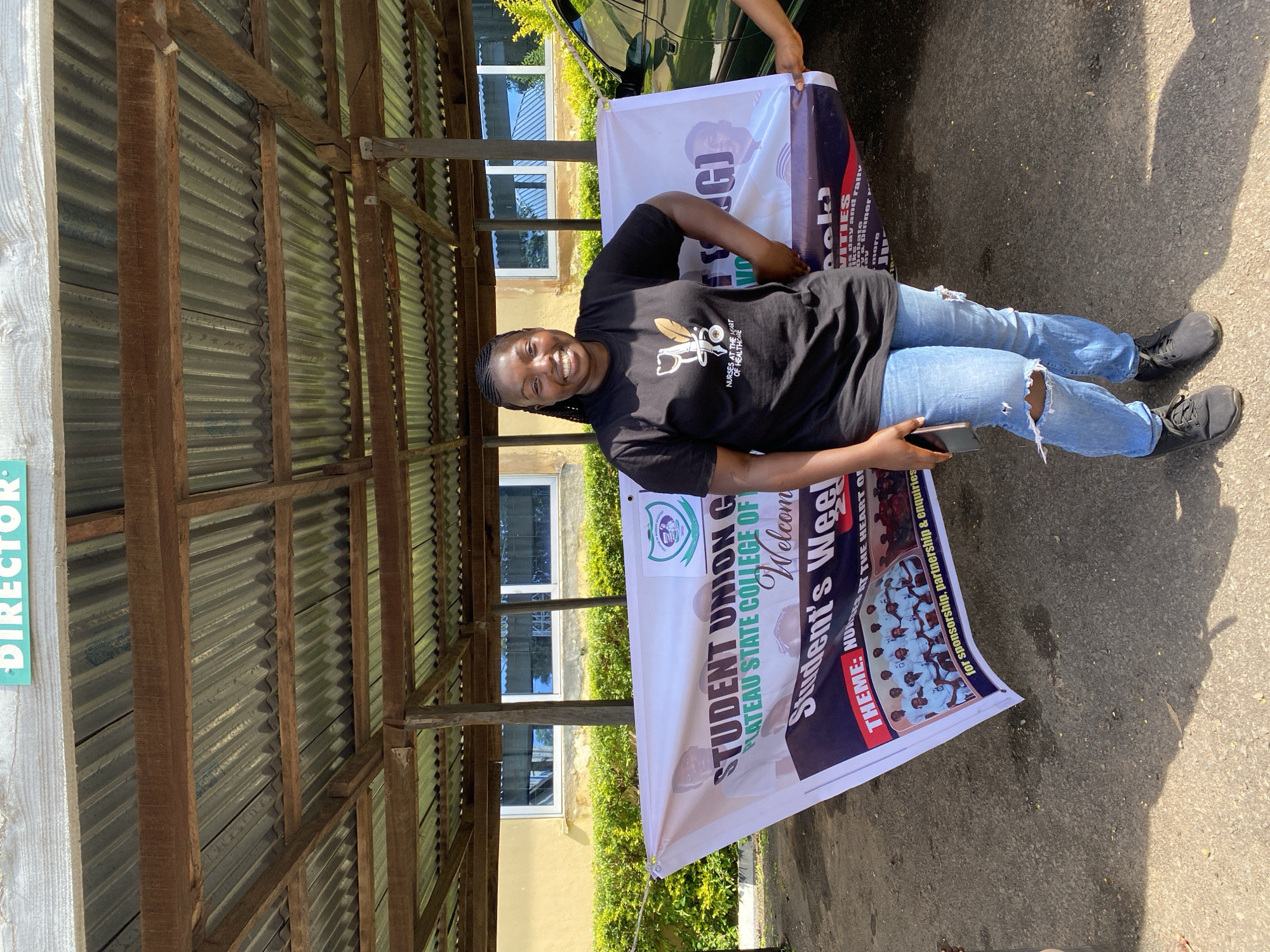 This media file captures a young Nigerian woman standing outdoors, holding a large banner. She is smiling broadly, exuding happiness and confidence. She is dressed casually in a black t-shirt with a stethoscope and feather logo, and ripped blue jeans. The banner, which she is holding by the corner, appears to be for a "Student Union Government" event at "Plateau State College of". Text on the banner indicates the event includes "Activities", a "Theme: Nurses at the heart of healthcare", and is for "sponsorship, partnership & enquiries."
The setting seems to be a covered outdoor area with a corrugated metal roof supported by wooden beams. Behind the woman, a building with windows and lush green foliage can be seen, suggesting a warm, possibly sunny day. The ground is asphalt, and shadows are cast on it, indicating the time of day is likely late morning or early afternoon. The presence of a vehicle, partially visible, further grounds the scene as an outdoor event. The overall impression is one of a cheerful and organized student event.