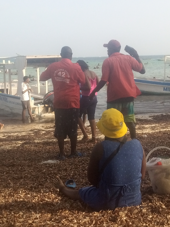 A daylight scene on a beach in Shanzu ward, Kenya, displays a shoreline covered in dried brown foliage, likely seaweed. In the foreground, a person with their back to the viewer sits barefoot on the foliage, wearing a yellow bucket hat and a blue patterned dress. A white plastic bucket with a handle is positioned to their right. Further back, near the water's edge, three individuals stand. A man on the left wears a red short-sleeved shirt with "KIMA LAKO STUDIO 42 IN PUBLIC BEACH PHOTOGRAPHERS" printed on the back and dark patterned shorts. Adjacent to him, a woman wears a pink tank top and dark shorts. To the right, another man in a red short-sleeved shirt, multi-colored shorts, and a red baseball cap holds a black object, possibly a camera or speaker, in his raised right hand. In the shallow water, a young male in a white t-shirt and dark shorts stands beside a white boat labeled "YANMAR" and "40 HP BOAT & SNORK," fitted with a Yamaha outboard motor. Multiple individuals are visible swimming or wading in the ocean further offshore.