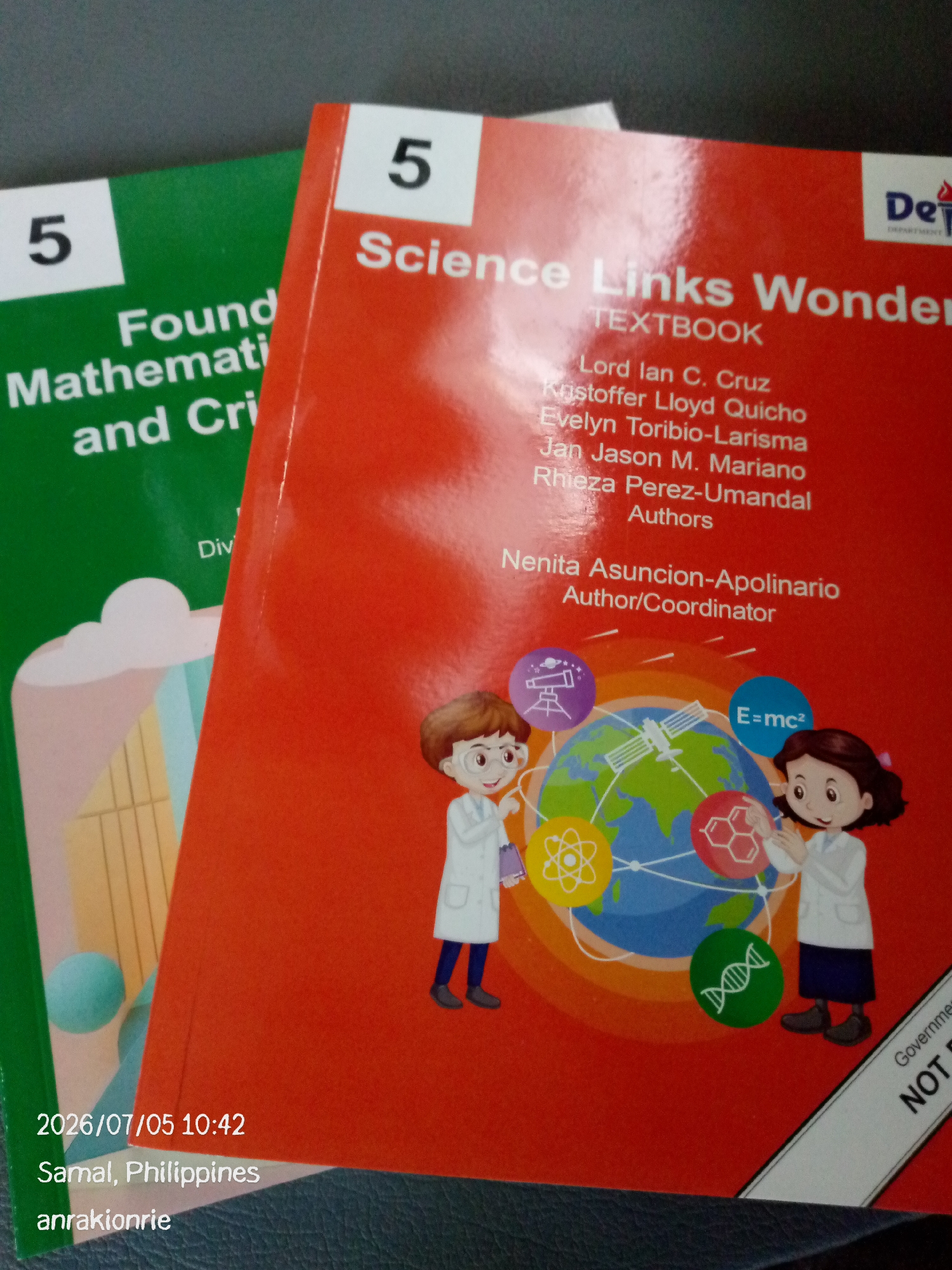 The image shows two textbooks. The textbook in the foreground is red with the title "Science Links Wonder" and "TEXTBOOK" below it. It also lists authors and an author/coordinator. There are illustrations of two children in lab coats interacting with a globe that has scientific icons on it, including a telescope, a DNA strand, and a molecular structure. The textbook in the background is green with the title "Found Math..." visible. The image is dated "2026/07/05 10:42" and located in "Samal, Philippines". There is also text at the bottom that reads "anrakionrie". A white diagonal strip with black text, partially obscured, reads "Government NOT P...". The lighting suggests it was taken indoors with some natural light.