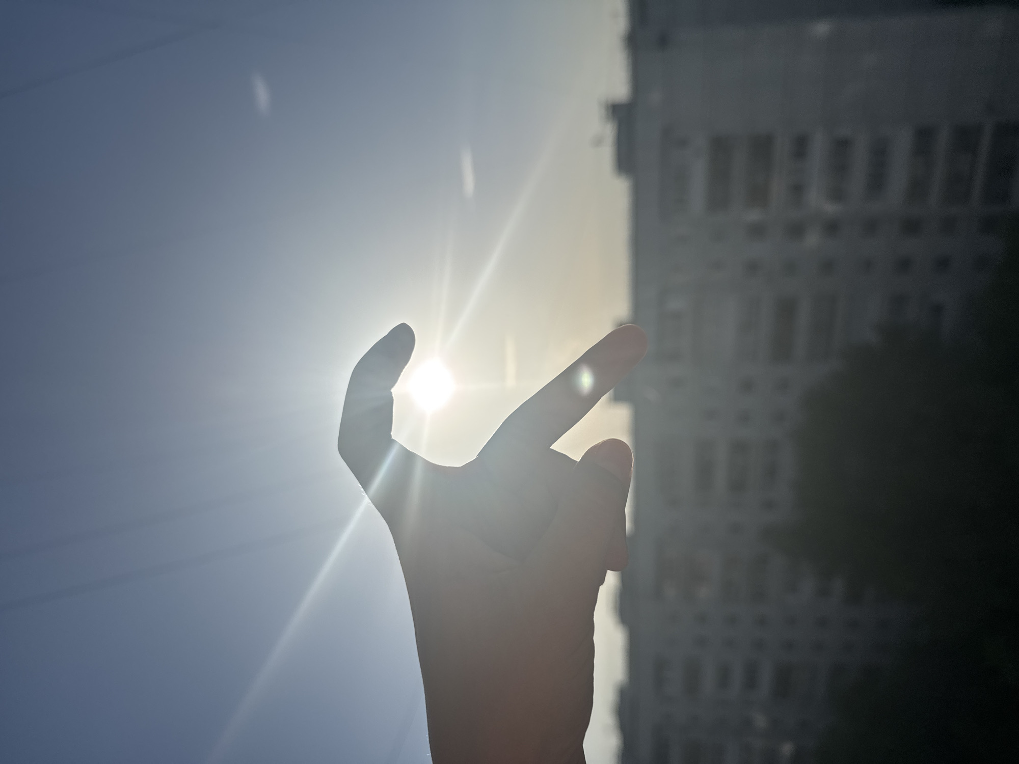 A hand is silhouetted against the sun, appearing to pinch or hold the sun between the thumb and index finger. In the background, a tall building with multiple windows can be seen. The sky is a gradient from light to dark. The specific location in Chaikovsky, Russia, cannot be determined from the image alone.