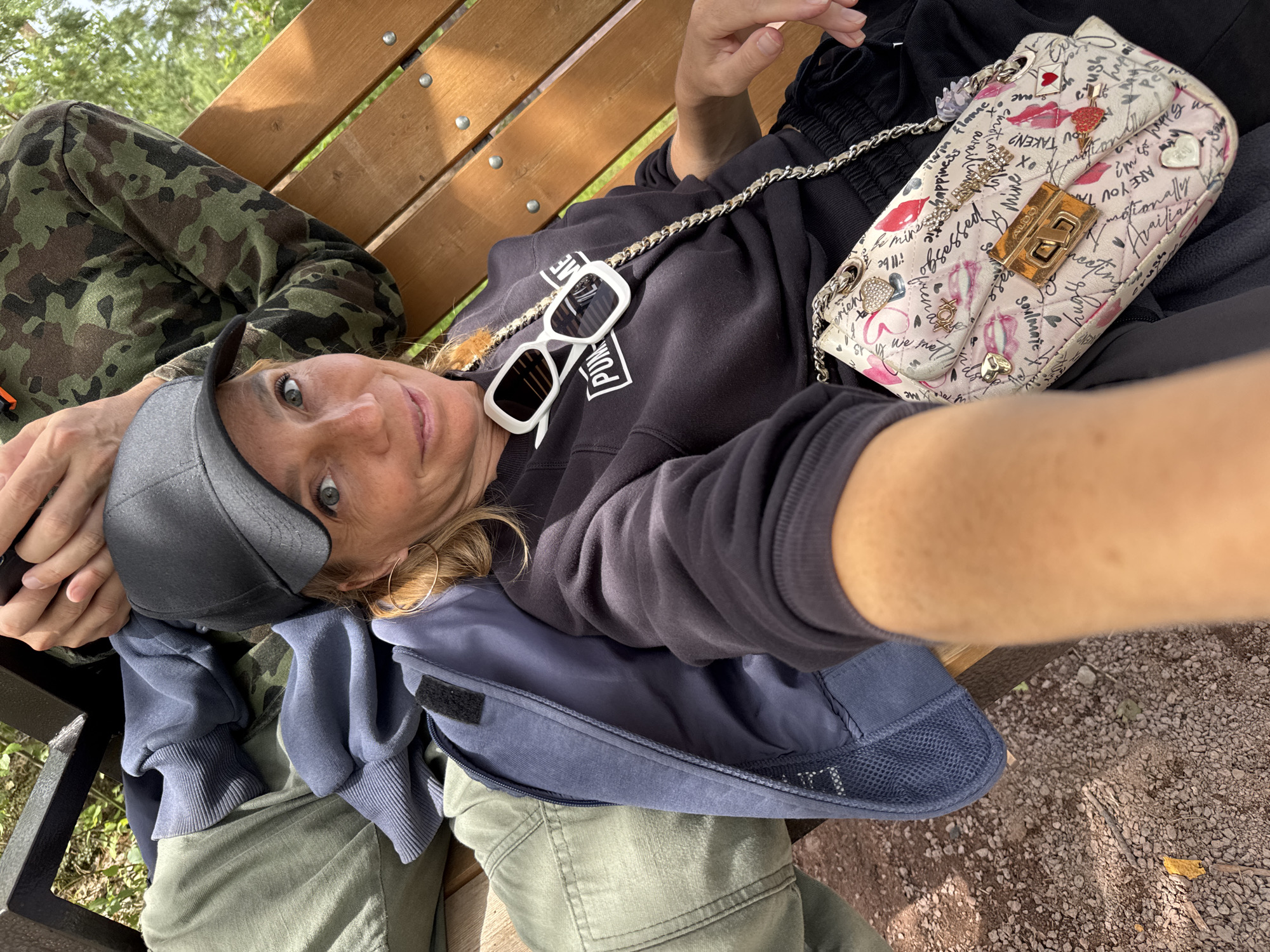 An outdoor, eye-level, medium shot captures two individuals sitting on a wooden park bench in Peski, Russia, with a woman in the foreground and another person partially visible in the background. The woman, positioned on the right, is wearing a black baseball cap, sunglasses on her head, and a black sweatshirt with a white logo. She is also accessorized with a crossbody bag adorned with various graphics and text. A person in camouflage clothing is seated to her left. The bench is made of wood with visible screws and is situated on a path covered with gravel and small stones. Sunlight filters through trees in the background, suggesting a daytime setting. The woman is looking directly at the camera, and the composition implies she is taking a selfie or being photographed from a close, personal angle.