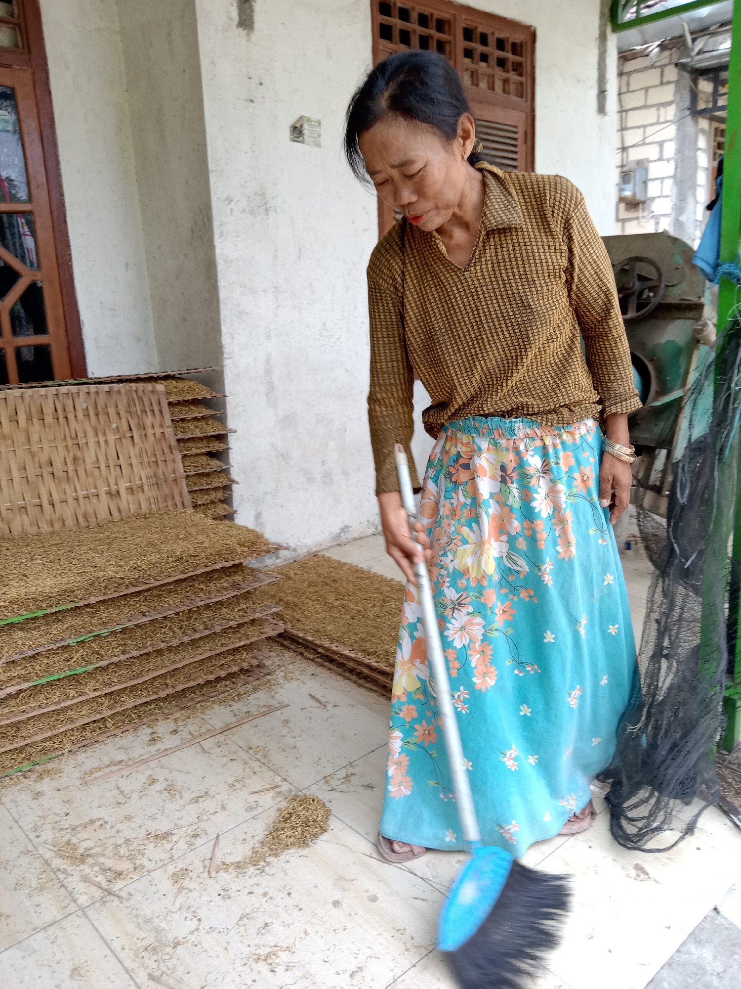 In a village setting in Jumput, Indonesia, a woman is sweeping the tiled floor. She is wearing a patterned long-sleeved shirt and a floral-patterned skirt. She is using a blue broom with a long handle to sweep debris. In the background, there are stacks of dried goods on woven mats, a wooden door and window, and some machinery. The scene appears to be an outdoor area adjacent to a building, possibly a dwelling or a workshop, with a glimpse of a green structure on the right.