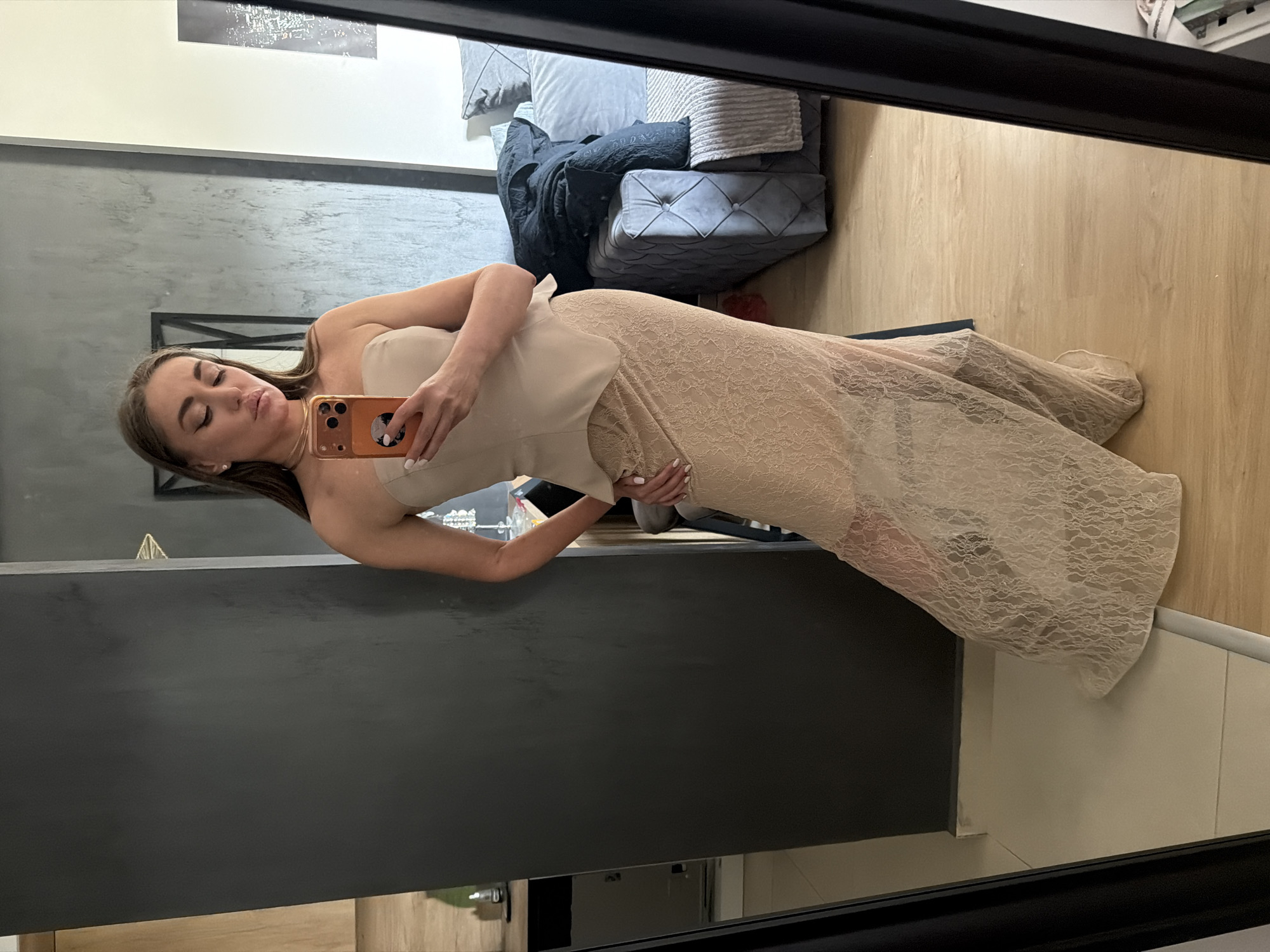 A young woman is captured in a mirror selfie, standing in what appears to be a modern indoor setting. She is wearing a strapless, light beige or nude-colored dress with a fitted bodice and a long, flowing skirt made of intricate lace. Her hair is long and brown, falling over her shoulders. She holds a light orange/gold smartphone with a pop socket or grip on the back, angling it to capture her reflection in the mirror. Her expression is relaxed, with slightly parted lips.
The background, visible through the mirror's reflection, shows a well-furnished room. To her left, there's a seating area with a gray sofa adorned with various cushions and blankets, and a tufted gray ottoman. The floor is covered with light wooden laminate. A dark, rectangular structure, possibly a counter or table, is prominent in the foreground of the reflection. The walls are a mix of light and dark gray, and a piece of artwork or a picture is visible on an upper wall. The overall ambiance suggests a contemporary living space, possibly an apartment or hotel room in Bar, Montenegro. The lighting indicates it is likely daytime. No visible text is discernible in the image.