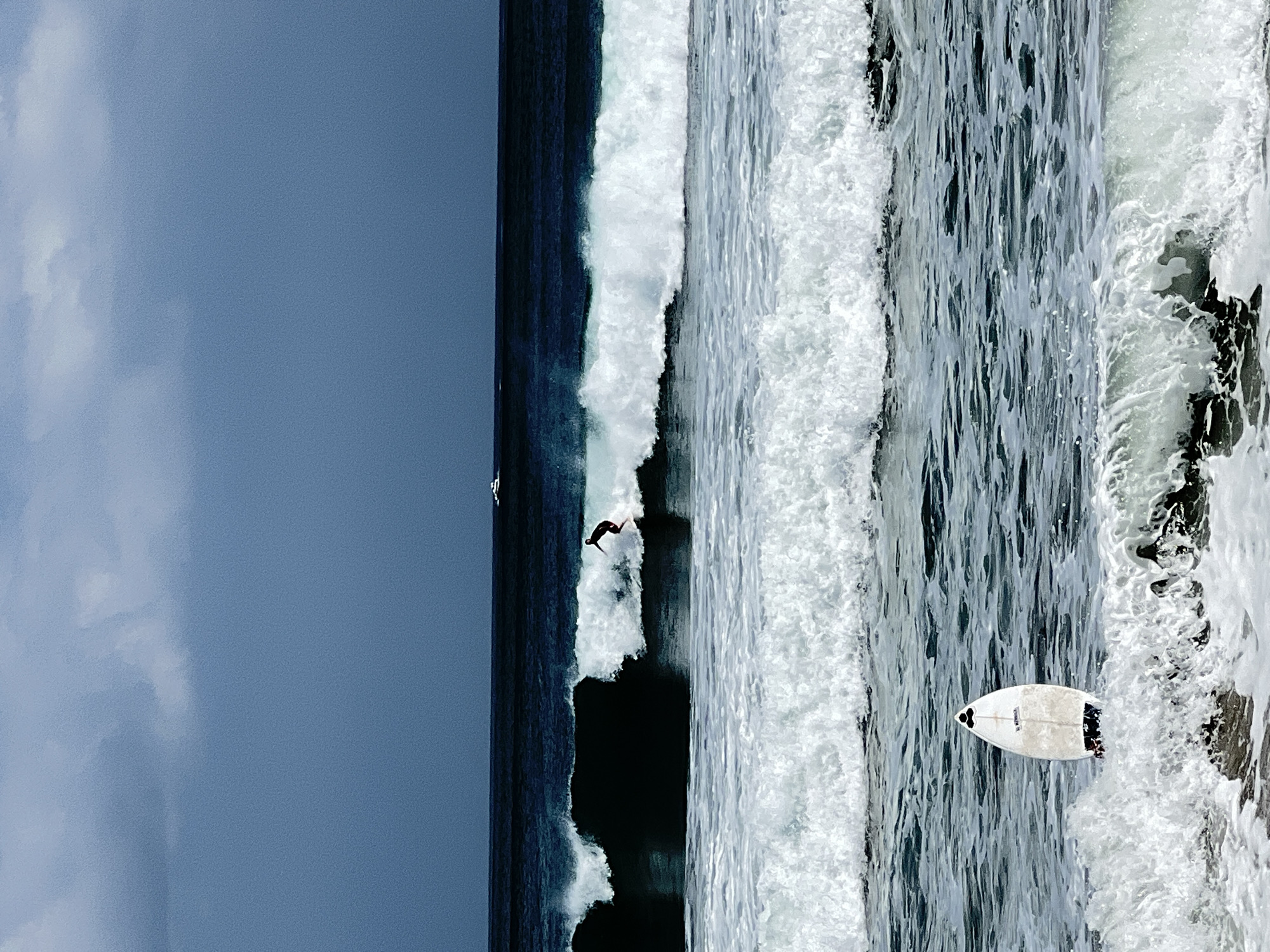 A surfer is riding a wave in the ocean under a cloudy sky. The surfer is a silhouette against the bright white foam of the wave. In the background, there is a small boat on the dark blue water. In the foreground, another surfboard is upright in the water, facing the camera. The ocean has multiple layers of waves, with white foam and dark blue water. The sky is a mix of dark and light blue clouds. The scene appears to be a popular surf spot, possibly in Kuta, Indonesia, known for its beaches and surf conditions.