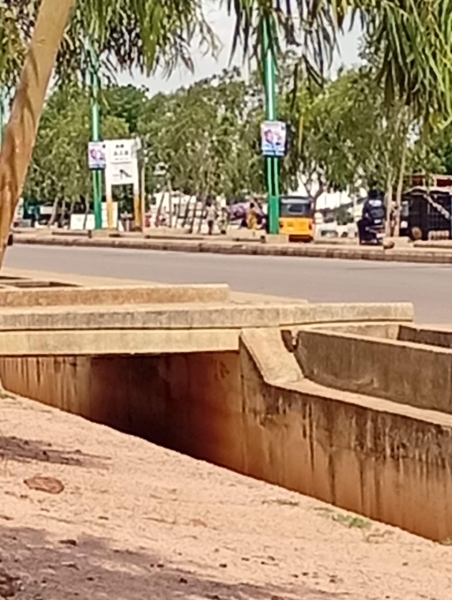 A medium shot captures a street scene in Tumfure, Nigeria. In the foreground, a concrete structure, possibly a bridge abutment or planter, forms the lower portion of the image. Beyond this structure, a paved road is visible, with a sidewalk bordering it. Several vehicles are present: a yellow auto-rickshaw is parked on the sidewalk, and to its right, a black car is stopped. People are present on the sidewalk. Behind the road, trees and foliage create a backdrop, and a street sign with a blue and white sign and a smaller poster is visible. The overall scene depicts a typical urban street environment.