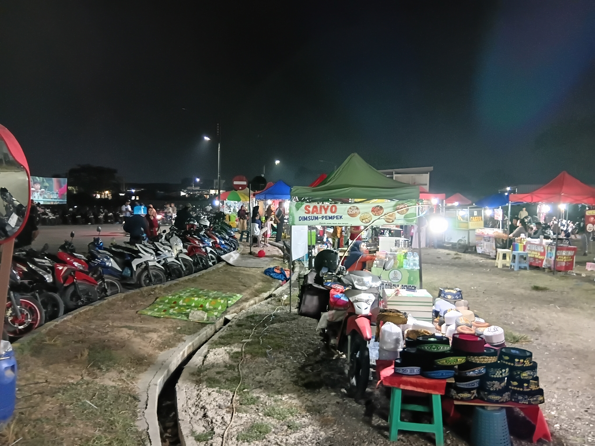 The image captures a night scene at a market in Sukasejati, Indonesia. The foreground features a variety of motorcycles parked along a curved concrete ditch. To the right of the motorcycles, a stall displays a collection of Islamic prayer caps, known as peci or kopiah, arranged on a red cloth-covered table and a green stool. Beyond the motorcycles and the cap stall, multiple market stalls with colorful tents are visible. One stall prominently displays a banner for "SAIYO DIMSUM-PEMPEK," indicating food vendors. People are seen browsing and interacting at the stalls. The environment appears to be an open, unpaved area, illuminated by artificial lighting from the stalls and streetlights, casting a warm glow on the scene. The sky above is dark, characteristic of nighttime.