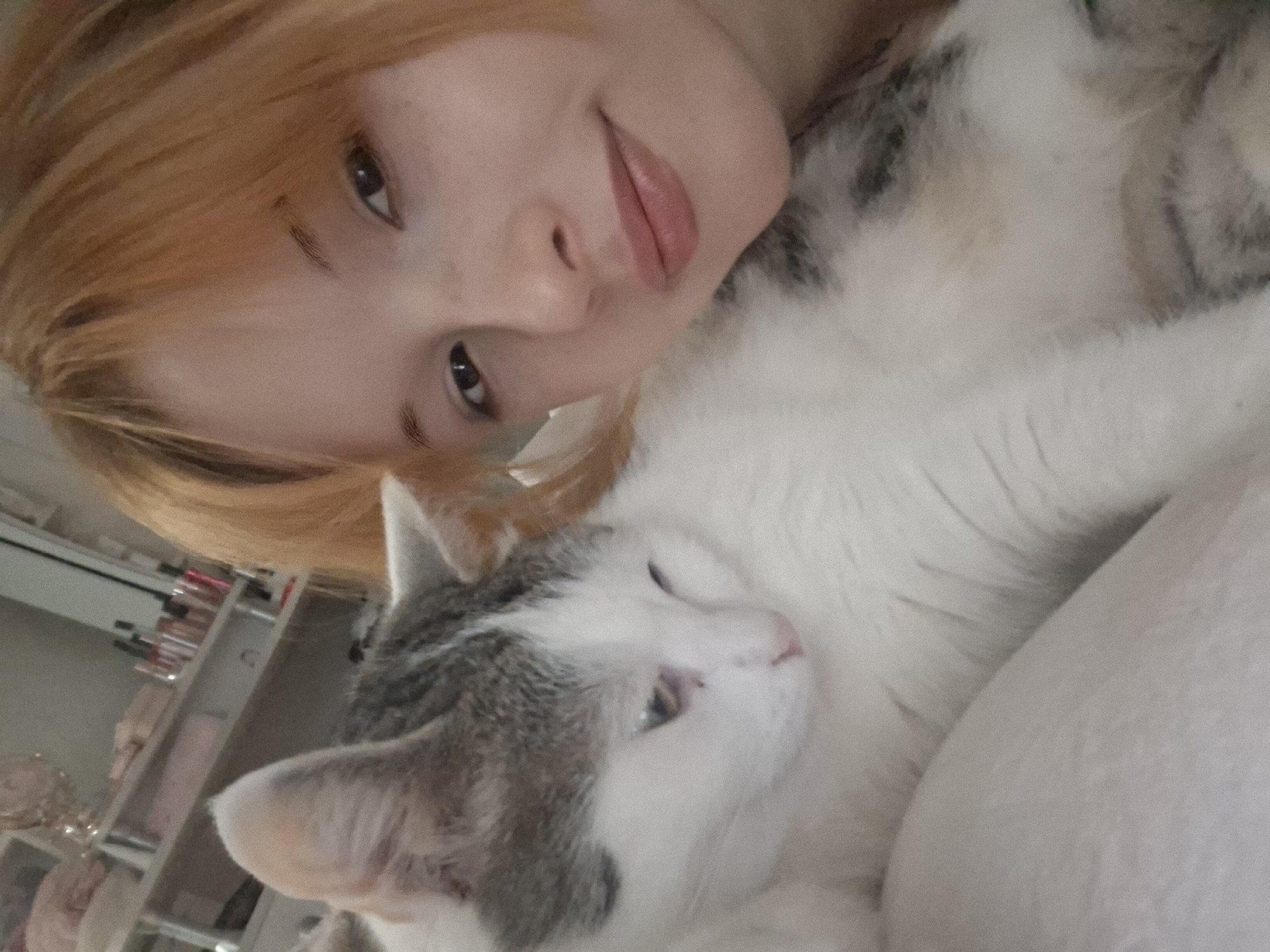 A young woman with ginger hair is pictured in a selfie with a gray and white cat. The woman has light skin, brown eyes, and is smiling slightly. The cat is nestled against her, with its head resting on her arm or chest. The background is blurry but appears to be a room with shelves filled with cosmetic or skincare products. There is no information in the image to confirm the location in the city of Chaikovsky, Russia.