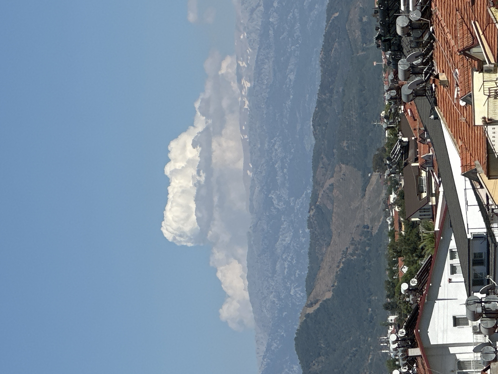 This image captures a panoramic view of a mountainous landscape overlooking a town or village, likely in Fethiye, Turkey. The scene is dominated by a clear, light blue sky with several bright white, puffy cumulus clouds scattered across the upper left and middle.
In the mid-ground, multiple layers of mountains stretch horizontally, receding into the distance. The mountains closer to the foreground appear greener and more detailed, while those further back are progressively hazier and bluer, indicating atmospheric perspective. Some lighter patches are visible on the distant mountain slopes, possibly bare rock or arid areas.
The foreground, primarily occupying the right side of the image, features numerous residential buildings with distinct red-tiled roofs. Many of these rooftops are heavily equipped with various installations, including multiple silver, cylindrical water tanks, an array of black rectangular solar panels, and numerous white satellite dishes. The architectural style suggests a Mediterranean influence, common in Turkey. Green trees and foliage are visible among the buildings and on the lower mountain slopes, adding to the natural setting.
The lighting indicates bright daylight, likely mid-morning or afternoon, given the clear sky and well-illuminated features. There is no visible human activity or text within the image. The overall impression is one of a serene, sunny day in a scenic, elevated location overlooking a populated area nestled within a mountainous region.