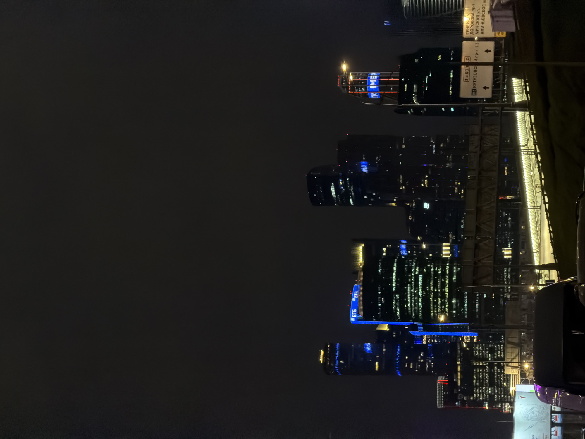 The image depicts a nighttime view of the Moscow International Business Center (Moscow City) skyline. High-rise buildings with illuminated windows and blue signage are visible against the dark sky. Road signs indicating directions to Kutuzovsky Prospekt, Dorokhova Prospekt, Minskaya Street, and Aminyevskoe Highway are prominently displayed. A bridge with railings is also visible. The scene is captured at night, with a focus on the urban environment and infrastructure.