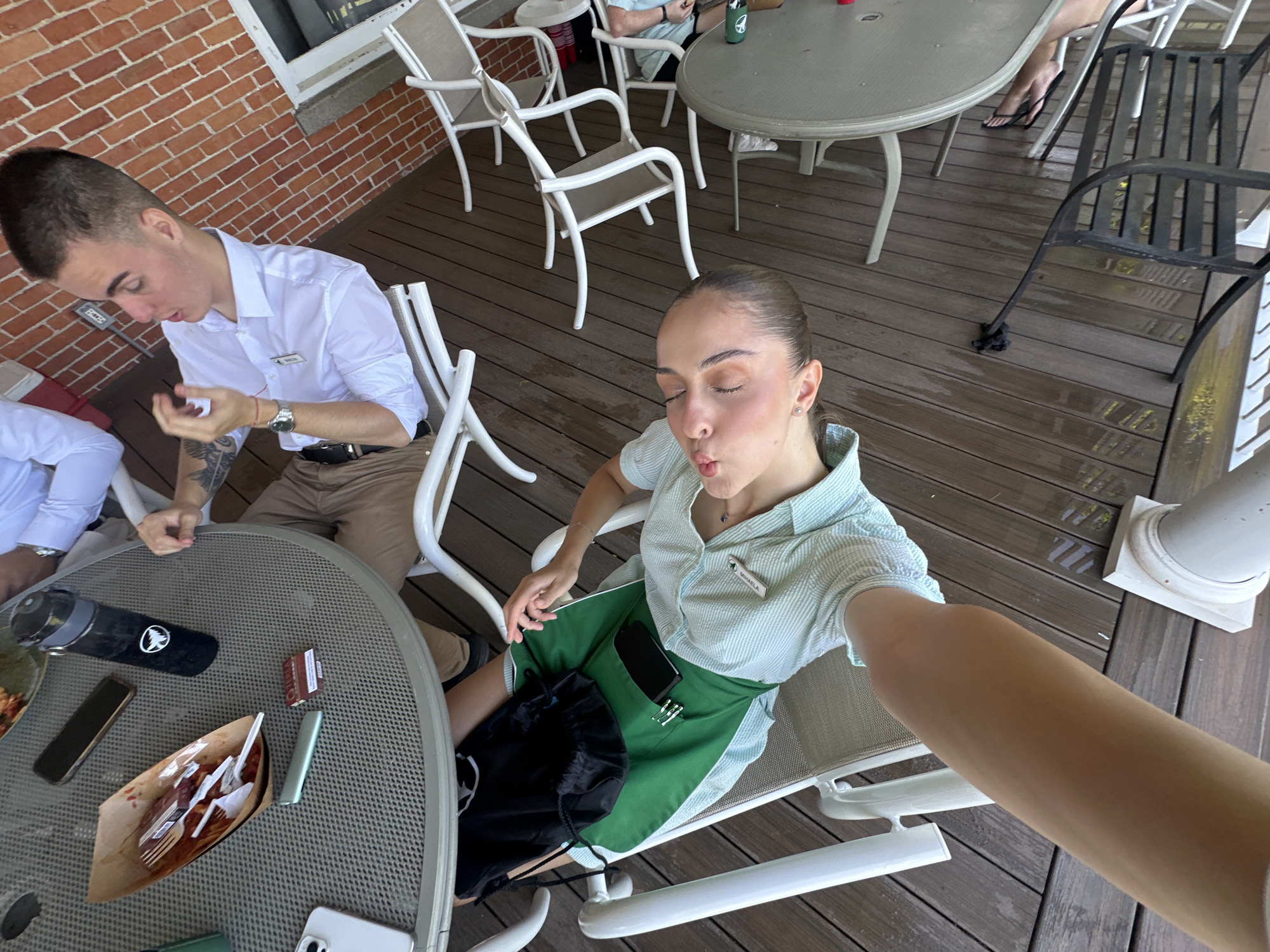 The image captures an outdoor gathering on a wooden deck. In the foreground, a person, possibly a woman, is lying down and taking a selfie, puckering her lips. She is wearing a light blue and white striped shirt and green shorts. A black drawstring bag rests on her lap.
In the background, a man in a white collared shirt and khaki pants is seated at a round table. He appears to be gesturing with his hand. The table has a brown paper container with utensils, a black water bottle, and a small rectangular object on it.
Several white outdoor chairs are scattered around the deck. A brick wall is visible in the upper right portion of the image. The wooden deck appears to be wet in some areas. The overall scene suggests a casual outdoor social event. The provided crops offer closer views of the table items and the individuals. The location is identified as Harbor Springs, United States of America.