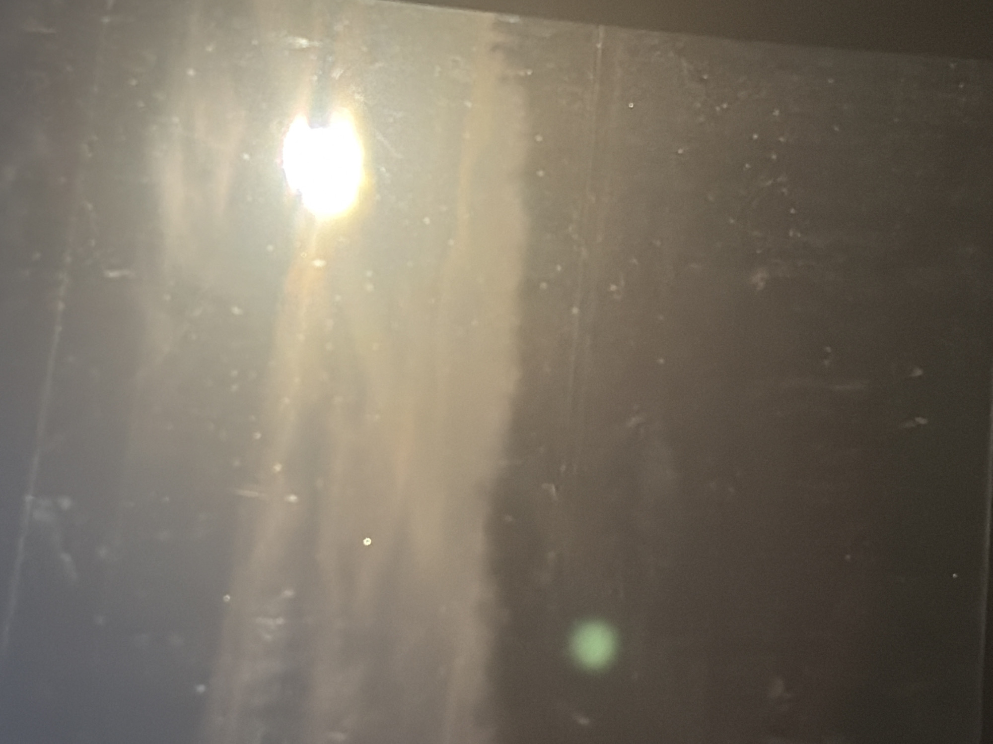 The image is a photograph taken through a window, likely from an indoor perspective looking outwards. The view depicts an outdoor scene with a bright, luminous object in the upper right quadrant, possibly the sun or a strong light source, casting a glow and creating lens flare effects. The sky appears hazy or overcast, with subtle cloud formations visible. In the mid-ground, there is a blurred landscape suggesting trees or a treeline. In the lower left quadrant, there is a distinct green circular light artifact, likely another lens flare or reflection. The window itself appears to have dust or smudges, contributing to the overall hazy and slightly distorted appearance of the image. There are no visible people, objects, or identifiable structures within the scene. The context provided about the location in Belogorki, Russia, cannot be verified from the visual information alone.