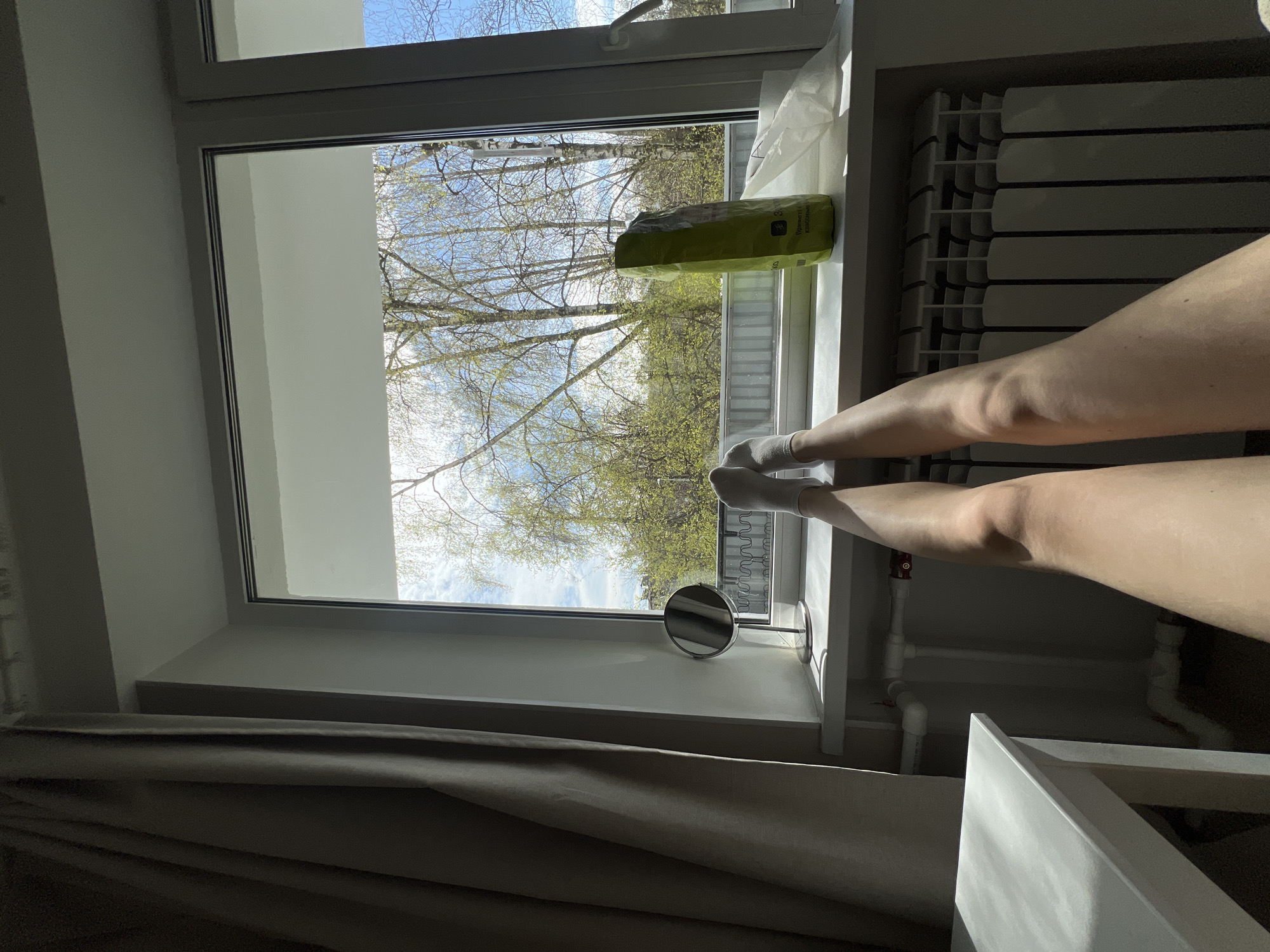 The image captures a first-person perspective from an interior room, looking out a window on a bright, sunny day. A person's bare legs, wearing white socks, are casually propped up on the white window sill, extending towards the right side of the frame.
Outside the window, the view reveals a clear blue sky with some scattered white clouds. Numerous deciduous trees, likely birches, are visible with their slender branches, just beginning to show the light green foliage of early spring.
On the white window sill, to the right of the person's legs, rests a light green, possibly foil, package. Russian text is visible on the package, including "Приметы" (Primetny, meaning "Signs") and "Порошкообразный" (Poroshkoobrazny, meaning "Powdered"), suggesting it contains a powdered product. A small, circular, silver-framed mirror with a stand is also positioned on the window sill, to the left of the legs.
Below the window sill on the right, a white radiator with horizontal panels is clearly visible. White pipes and a red valve, part of the heating system, are also present in the lower right corner, partially obscured by the window structure. Dark grey curtains are drawn partially across the lower left part of the window, creating shadows within the room. The bright sunlight streaming in creates strong contrasts and highlights the scene, suggesting a peaceful, relaxed moment during daytime in Saint Petersburg, Russia.
