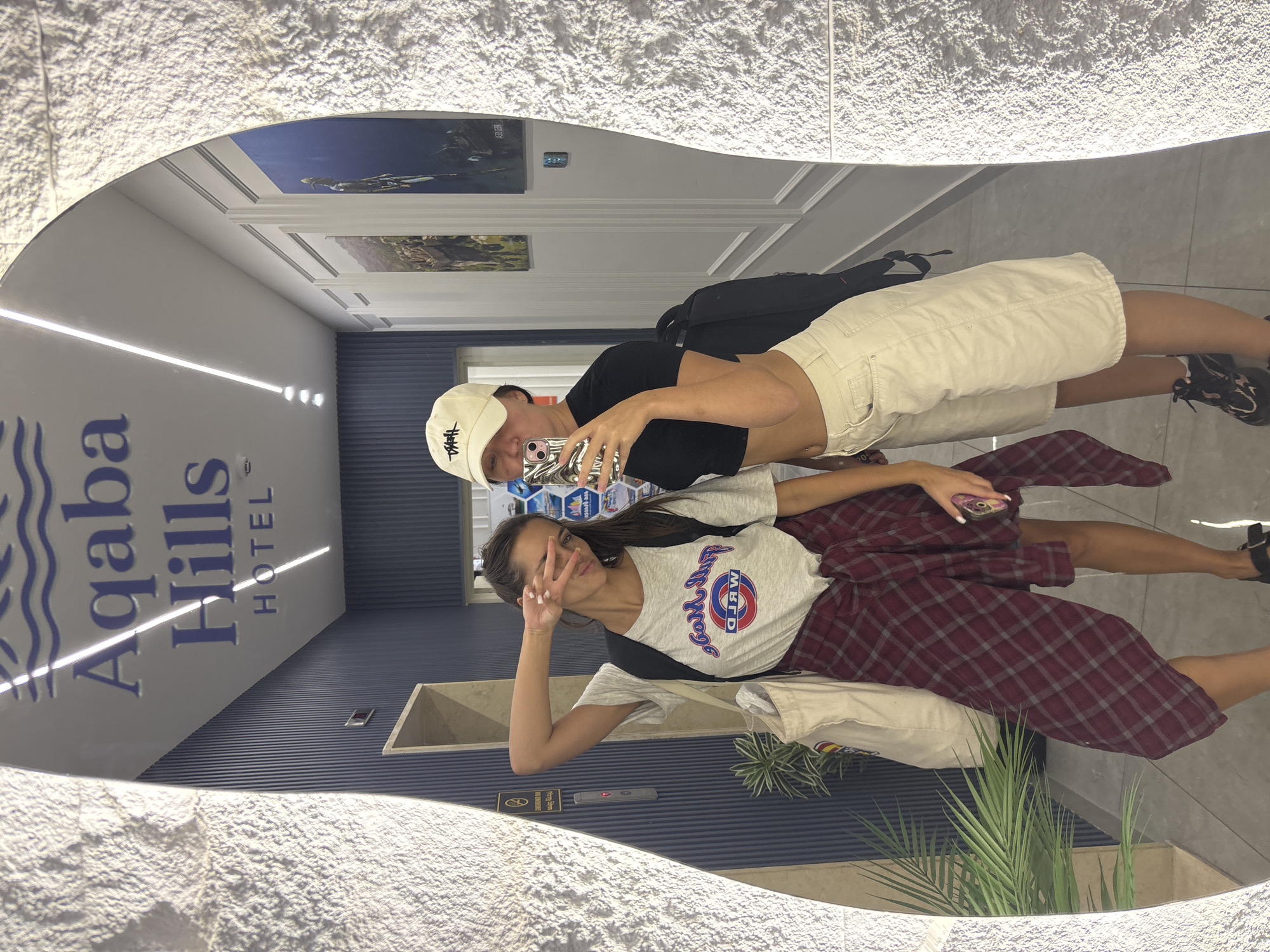 Two young women are taking a mirror selfie in a hotel hallway. The wall behind them features the "Aqaba Hills HOTEL" logo. The woman on the left is wearing a grey t-shirt with a red and white logo, a maroon plaid shirt tied around her waist, and black sandals. She has her left arm raised and is making a peace sign with her fingers. The woman on the right is wearing a black crop top, a white baseball cap, and beige shorts. She is holding a smartphone and taking the picture. A black backpack is visible behind her. The hallway is decorated with framed artwork on the walls and a potted plant.