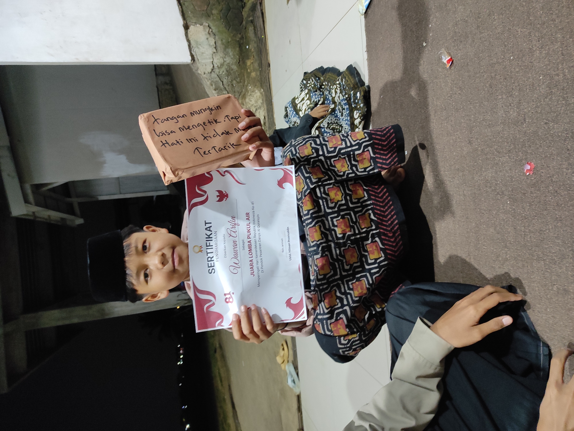 A young boy wearing a black peci hat holds a certificate and a gift. He is seated on a carpeted floor, likely outdoors at night, given the dark background with blurred lights. The certificate, titled "SERTIFIKAT PENGHARGAAN," indicates he is "JUARA LOMBA PUUKUL AIR" (Champion of the Water Pounding Competition), commemorating Indonesia's Independence Day at Pondok Pesantren Darqis Al Quraisyin. The gift is a brown paper-wrapped box with handwritten text in Indonesian. The boy's left hand is slightly visible, and his right hand holds the certificate. To the left, the arm and lap of another person in a light-colored shirt are visible. To the right, another person is seated in the background, wearing a patterned sarong. The environment appears to be an outdoor gathering space, possibly a courtyard or common area, with a concrete structure visible in the background and scattered debris on the floor. The location is specified as the city of Sumedang, Indonesia.
