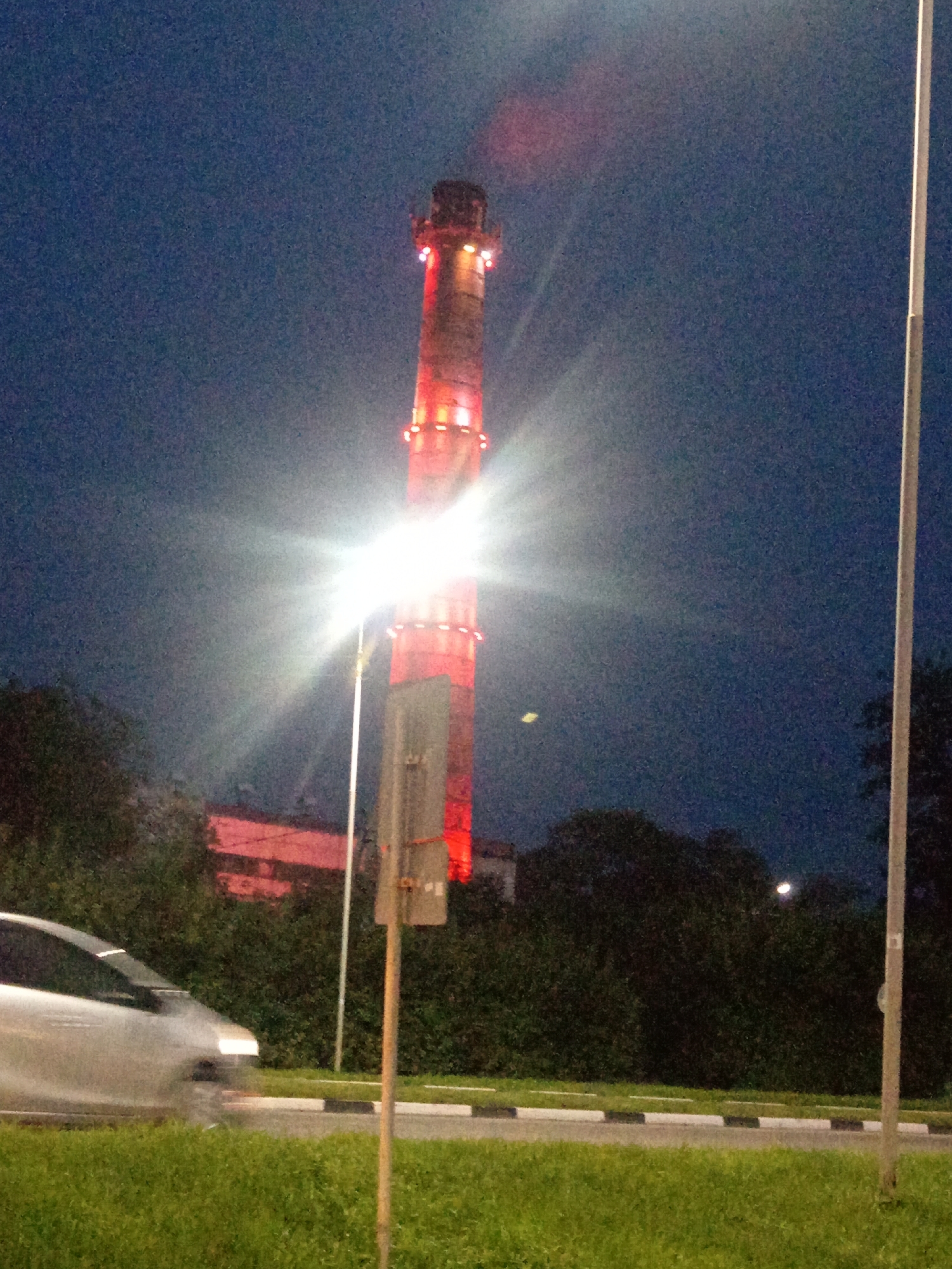 The image captures a nighttime view of a tall, cylindrical industrial chimney prominently lit with red and white lights, standing against a dark blue sky. A bright light source, possibly a streetlamp or floodlight, emanates intensely from the lower-middle section of the frame, creating a strong glare that obscures details in its immediate vicinity and casts light rays across the sky. The chimney appears to be a functional structure, possibly part of a power plant or factory. In the foreground, a silver car is captured in motion, blurred by its speed, driving on a road. The road is bordered by a grassy verge and a sidewalk marked with alternating black and white stripes. Behind the road and car, dense foliage of trees and bushes forms a dark silhouette against the illuminated chimney and the sky. Partially visible in the background, behind the trees, are the red-lit structures of other industrial buildings. A thin, vertical white pole, likely a street sign or utility pole, stands to the right of the chimney, and another pole is visible slightly to the left of the chimney, partially obscured by the glare. The overall scene suggests an industrial area at night, with the chimney being the most striking and illuminated element. The presence of a car and road indicates a populated or accessible area. The location is identified as Belogorsk, Russia.
