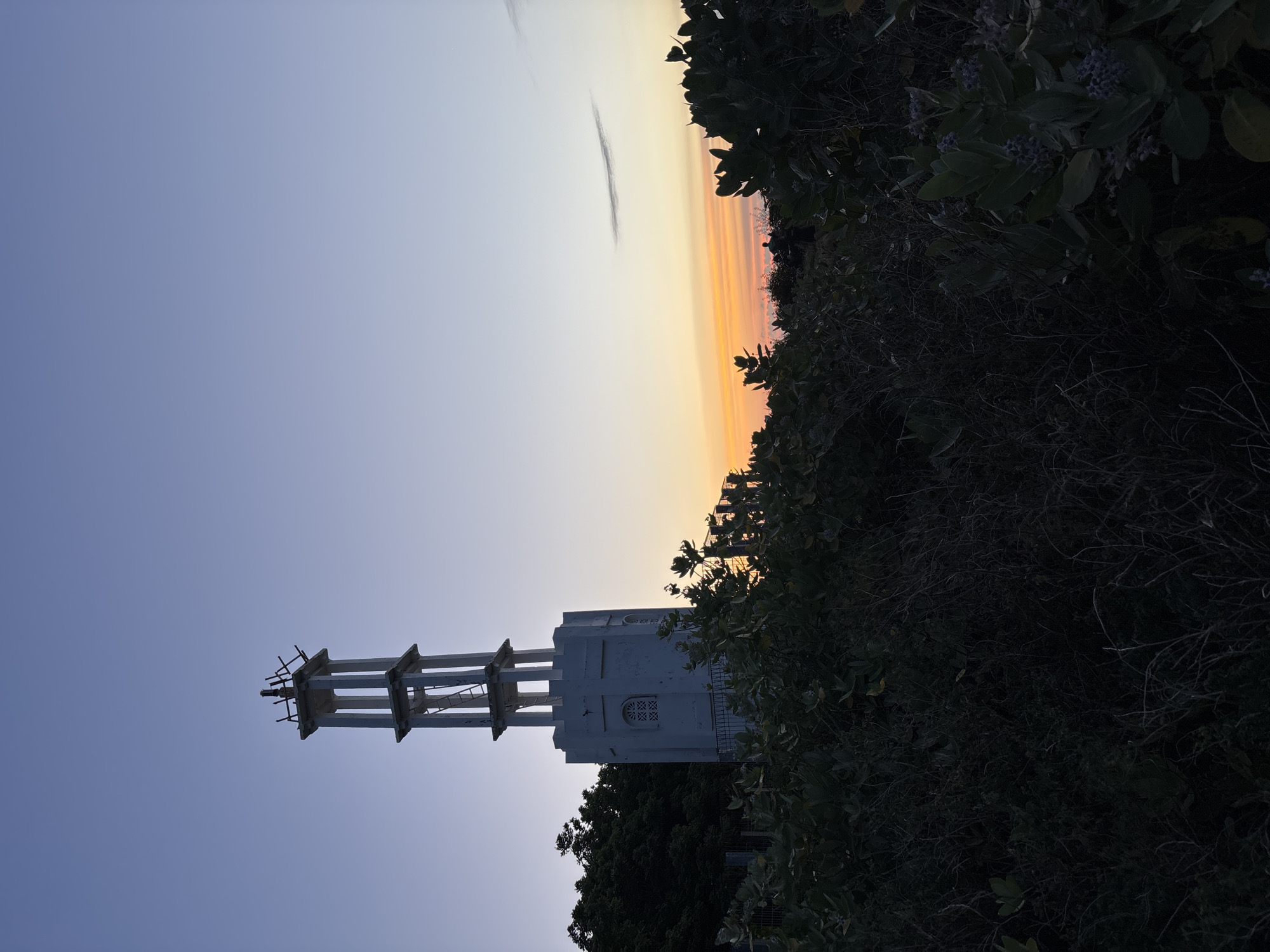 The image is an eye-level shot of a white tower or monument with a sky and sunset backdrop. The sky transitions from a light blue to an orange glow near the horizon. The tower is composed of a rectangular structure with vertical and horizontal beam supports, and what appear to be windows on the side. Vegetation, including leafy plants and possibly trees, frames the right side of the image. The perspective suggests the photograph was taken from a location in Pecatu, Indonesia. There are no visible people.