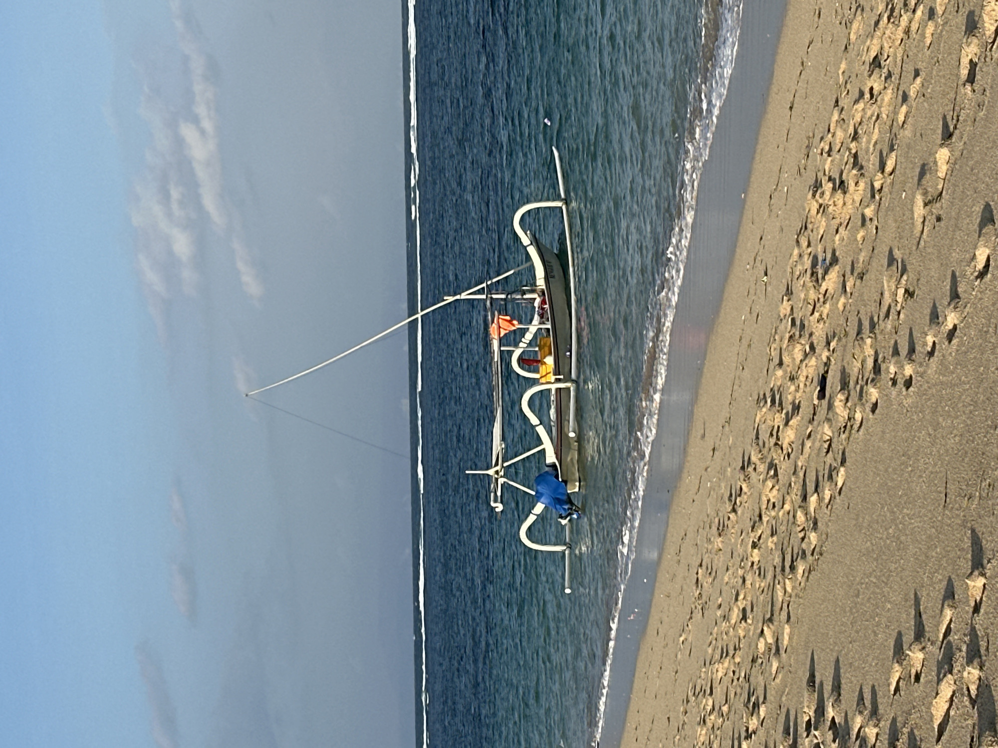A traditional Indonesian outrigger fishing boat (jukung) is positioned in the shallow, clear turquoise waters of a beach in Kuta Selatan, Indonesia. The foreground displays light brown sand covered with numerous footprints and minimal debris, where it meets the gentle white foam of an incoming wave. The jukung features a light-colored hull, distinctive white curved outriggers (cadik) extending laterally, and a tall, slender white mast angled slightly towards the stern. A blue tarp or bag is stowed on the port side of the boat, while a yellow bucket and an orange life vest are visible on the starboard side near the stern. The water around the boat is transparent, revealing the sandy seabed. In the background, the ocean transitions to a deeper blue, marked by several distinct lines of white breaking waves further from the shore. The sky overhead is a clear light blue, interspersed with scattered, wispy white clouds. No individuals are present in the frame.