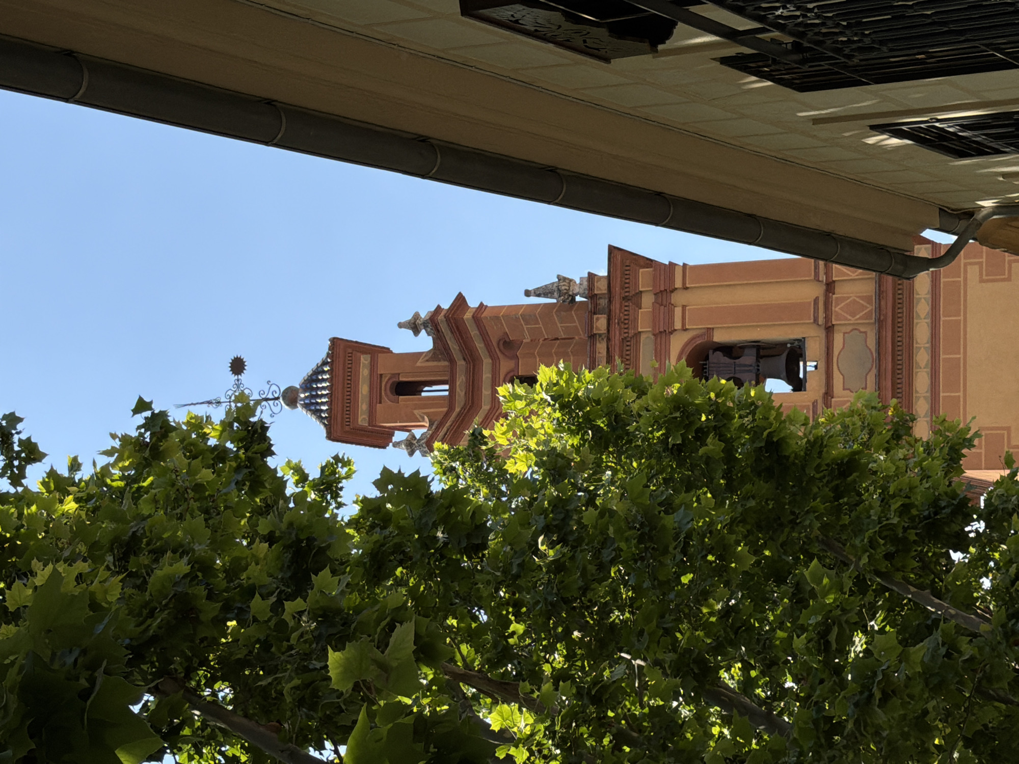 The image shows a close-up view of a bell tower, partially obscured by lush green foliage from a tree in the foreground. The bell tower is constructed of warm-toned masonry with decorative elements and features a bell visible within an arched opening. To the right of the tower, a portion of a building with a light-colored facade and a dark gutter system is visible. The sky above is clear and blue.