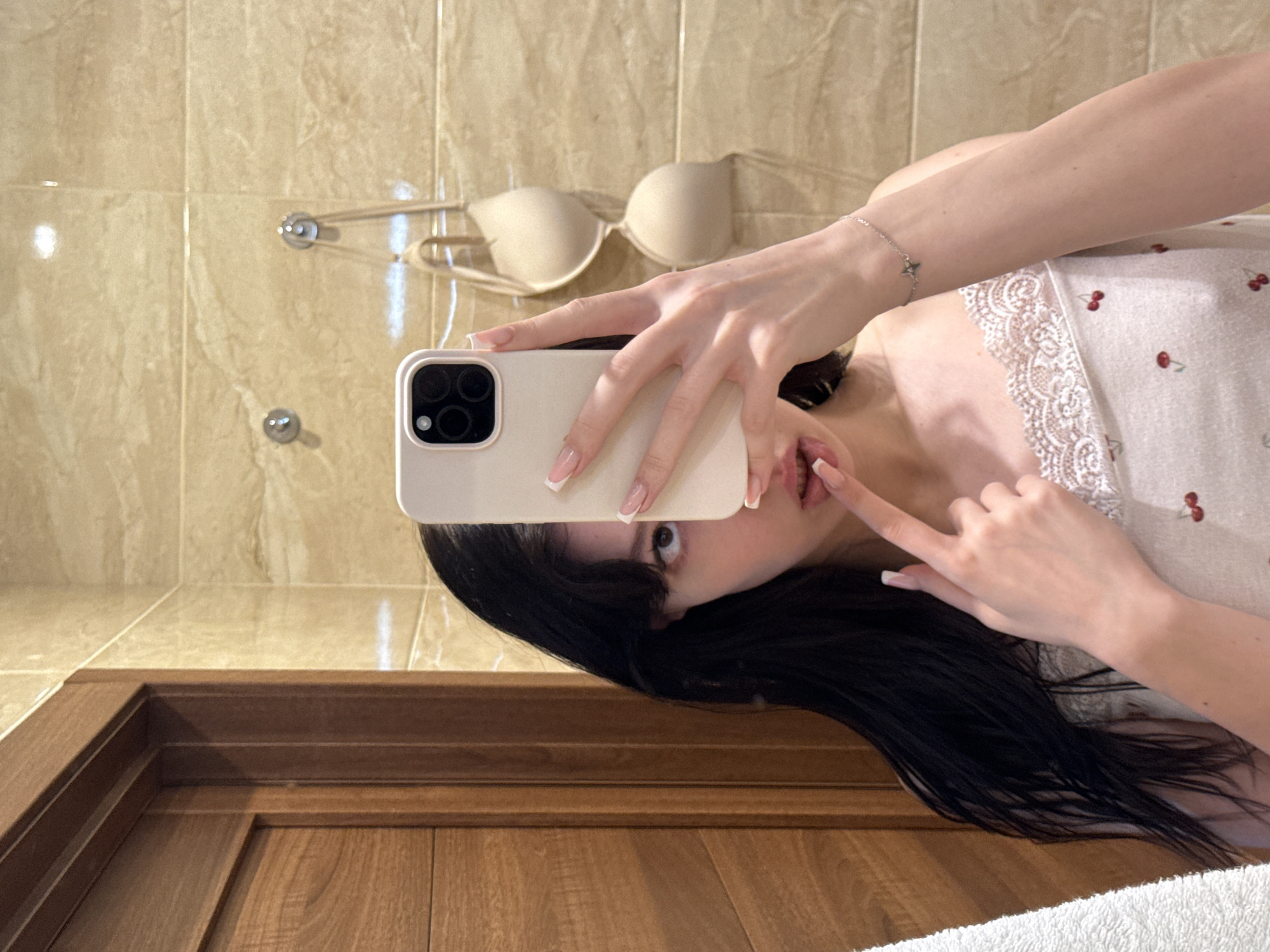 This is a selfie taken in a bathroom. A young woman with long, dark hair is holding a white smartphone to her face, obscuring part of it. She is wearing a white tank top with a lace trim and a cherry print. Her right hand is touching her lips, and her left hand is holding the phone. In the background, there are beige tiled walls with a wooden door on the left and a bra hanging on a hook on the right. There is a white towel on the far left. The image appears to have been taken in a private residence.