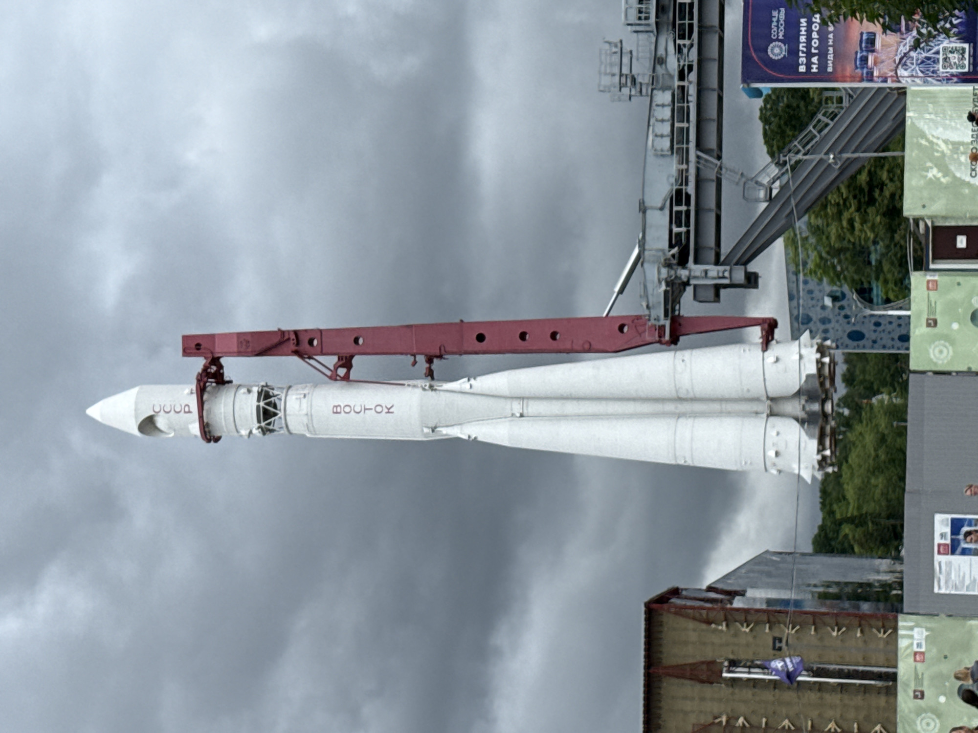 This media file features a Vostok rocket on display, likely in Moscow, Russia, given the visible text. The rocket, a historical artifact of the Soviet space program, is positioned vertically on a launch gantry or display structure. The rocket is predominantly white with red markings, including the word "CCCP" (USSR) and "BOCTOK" (Vostok) on its exterior.
The background is dominated by a cloudy, overcast sky, suggesting a cool or possibly rainy day. To the right of the rocket, a tall, multi-story structure is visible, with parts of it appearing to be a museum or exhibition space. Several posters or informational panels are attached to this structure. One prominent poster, oriented vertically, has text in Russian, including "СОЛНЦЕ МОСКВЫ" (Sun of Moscow) and "ВЗГЛЯНИ НА ГОРОД" (Look at the City). Another poster with the text "СКОТО ЗДЕСЬ БУДЕТ" (What will be here) is also visible.
While the focus is on the rocket, there are hints of people at the bottom right of the frame, suggesting a public viewing area. The overall atmosphere is one of historical significance and public interest in space exploration, set against a somber, cloudy sky. The scene appears to be outdoors, possibly in a park or open-air museum dedicated to space technology.
