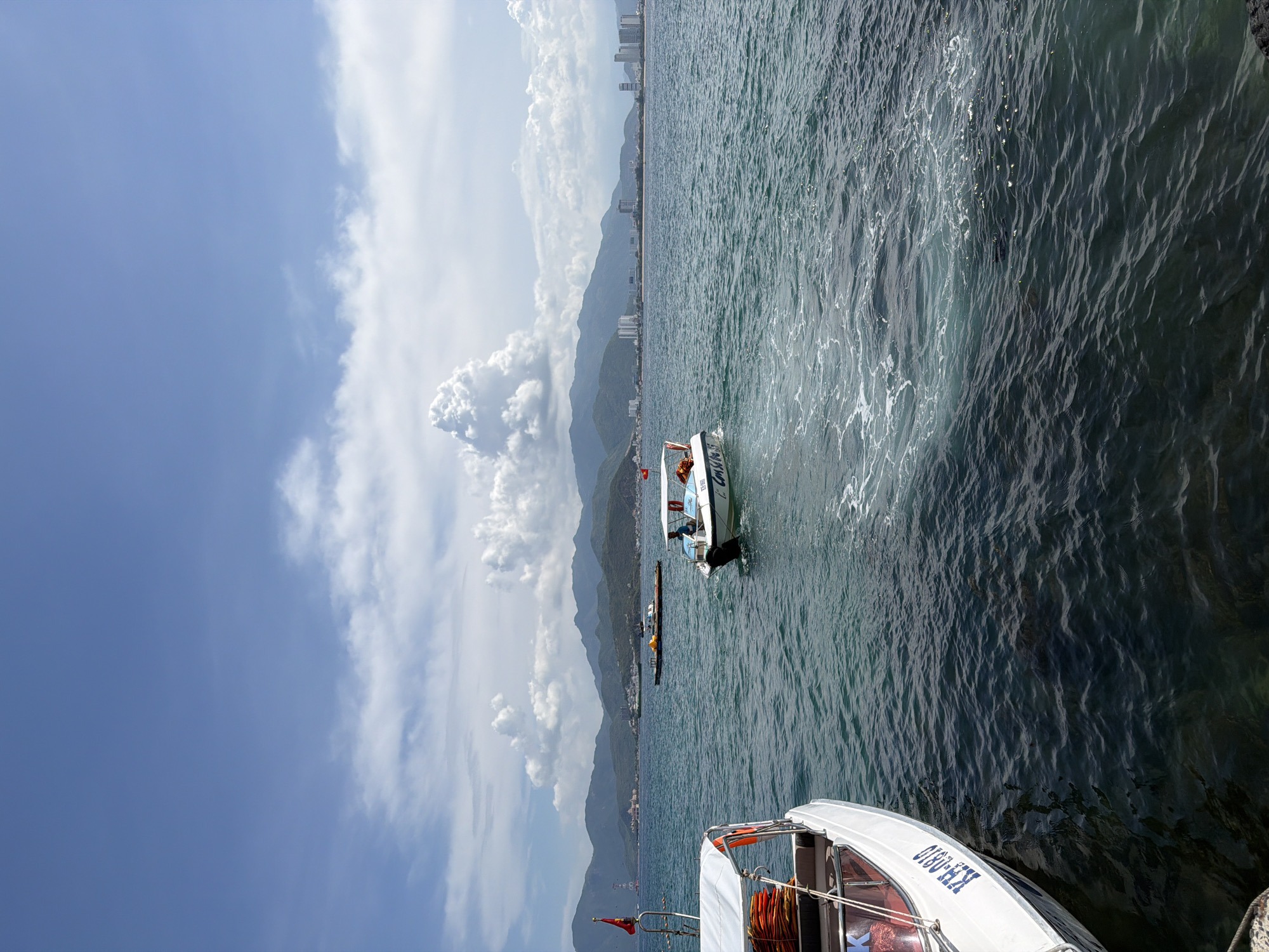 The image depicts a coastal scene in Nha Trang Ward, Vietnam. The sky is partly cloudy, with a gradient of blue sky near the horizon. A range of mountains runs along the coastline in the mid-ground, and high-rise buildings are faintly visible in the distance. Two small motorboats are in the water. The first boat is in the foreground and partially cut off by the frame, showing an orange life preserver and a Vietnamese flag. A second boat is visible mid-frame, with two people on board, trailing a visible wake. The sea appears calm but with ripples. There are three buoys aligned in the water. The angle of the photograph is looking downward.