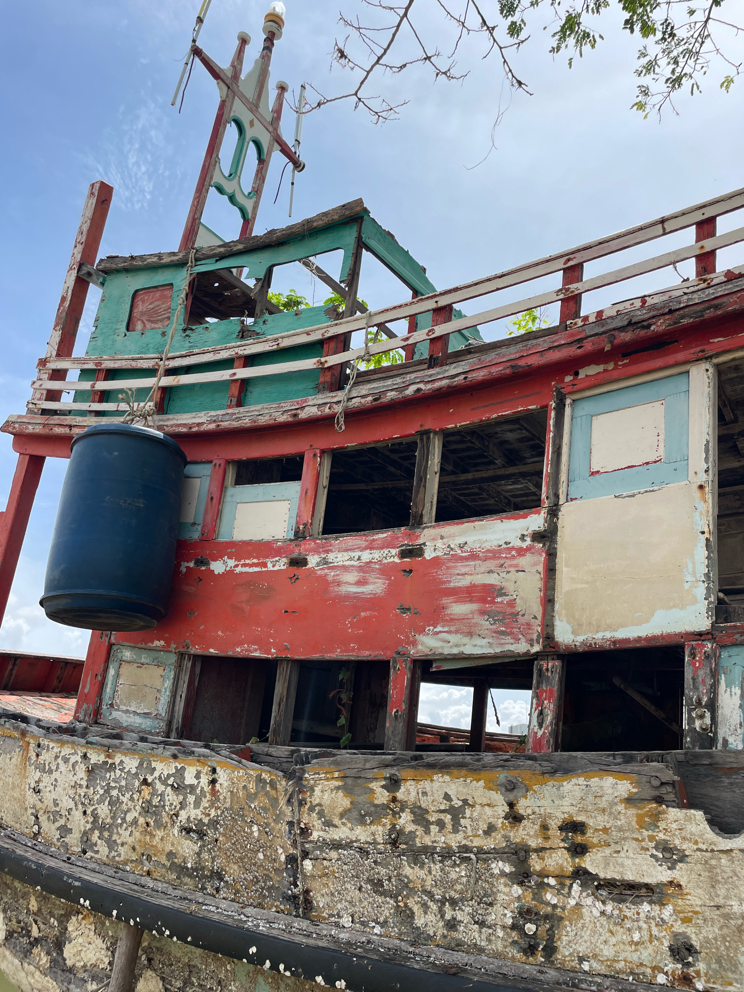 The image shows the dilapidated hull of a wooden fishing boat beached on the shore in Ban Na Chom Thian, Thailand. The boat's exterior is weathered and peeling, with layers of red, green, and white paint showing signs of decay. Barnacles and other marine growth encrust the lower sections of the hull. The upper structure of the boat is partially collapsed and appears to be made of wood, with broken railings and open windows revealing the interior. A large blue barrel hangs from a rope on the side of the boat. The sky is overcast with some clouds visible, and a few tree branches extend into the frame from the top right. The scene suggests the boat is abandoned and has been exposed to the elements for some time.