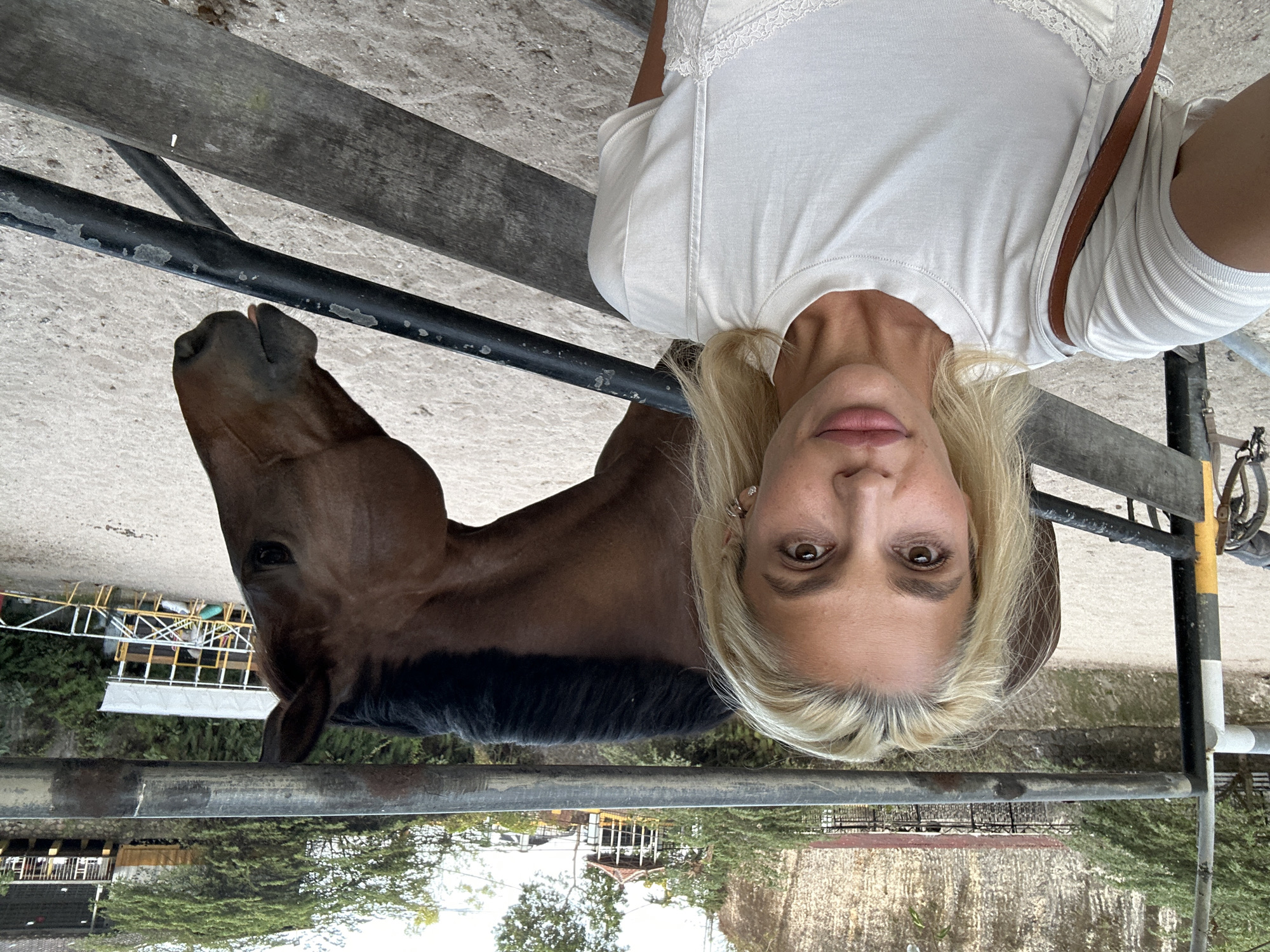 A fair-skinned woman, appearing to be in her 30s or 40s with blonde hair, light-colored eyes, and wearing a light-colored short-sleeved t-shirt over a lace-trimmed camisole, takes a selfie. A brown strap is visible over her right shoulder. She wears small earrings and light pink lip gloss. Directly behind her, separated by a metal and wooden fence, is a bay or dark brown horse, visible from its head and neck. The horse's head is turned slightly to its right, with its mane visible. The scene is an outdoor equestrian facility in Kuta Selatan, Indonesia. The ground is light-colored, sandy, or dusty. In the background, a large, textured rock face or cliff is prominent, with green trees and lush foliage growing on and around it. Several distant structures are visible through the trees, including a small gazebo with a reddish roof, and other light-colored shelters or buildings, characteristic of an equestrian arena or stable complex. Fencing consisting of metal bars and wooden rails separates the immediate foreground from the background subjects.