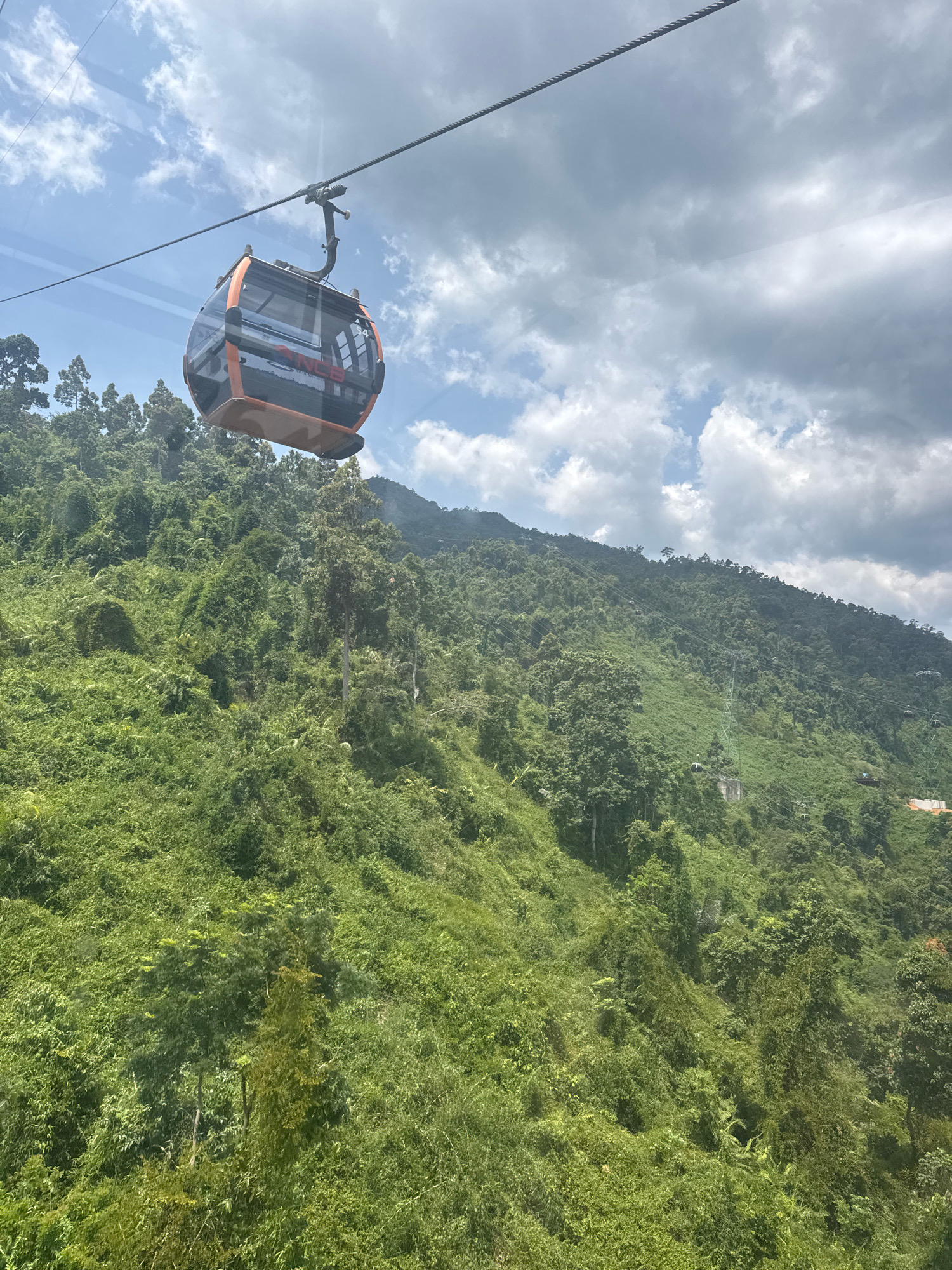 A cable car traverses a lush, mountainous landscape under a cloudy sky. The cable car, a prominent orange and black structure, is suspended from a thick cable, casting a shadow on the dense, green foliage below. The mountainside is covered in thick vegetation, with a mix of trees and smaller plants creating a vibrant tapestry of green. The sky above is partly cloudy, with patches of blue peeking through. In the distance, several other cable car towers and cabins are visible, suggesting a larger cable car system operating in the area. The overall scene depicts a scenic view of a natural landscape accessible by cable car.