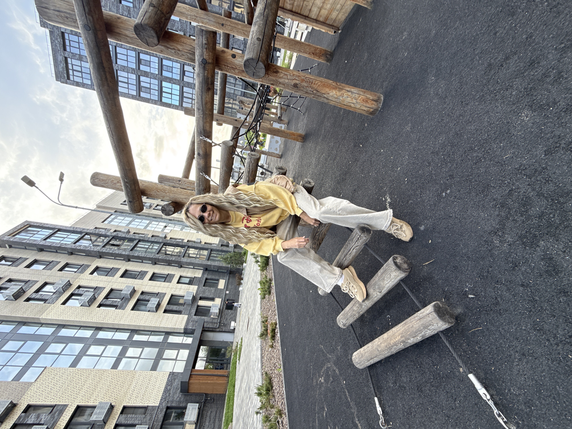 A full-length shot captures a fair-skinned, blonde-haired woman with sunglasses, seated on a wooden ladder structure in an outdoor area. She is dressed in a yellow sweatshirt, light-colored pants, and shoes. The ladder is constructed from wooden logs and suspended by ropes above a black, rubberized surface.
Behind the woman, the frame reveals a tall, modern building with numerous windows and a mix of light and dark gray brickwork. The environment is further characterized by some greenery near the building and a cloudy sky.
The location is an urban outdoor space.