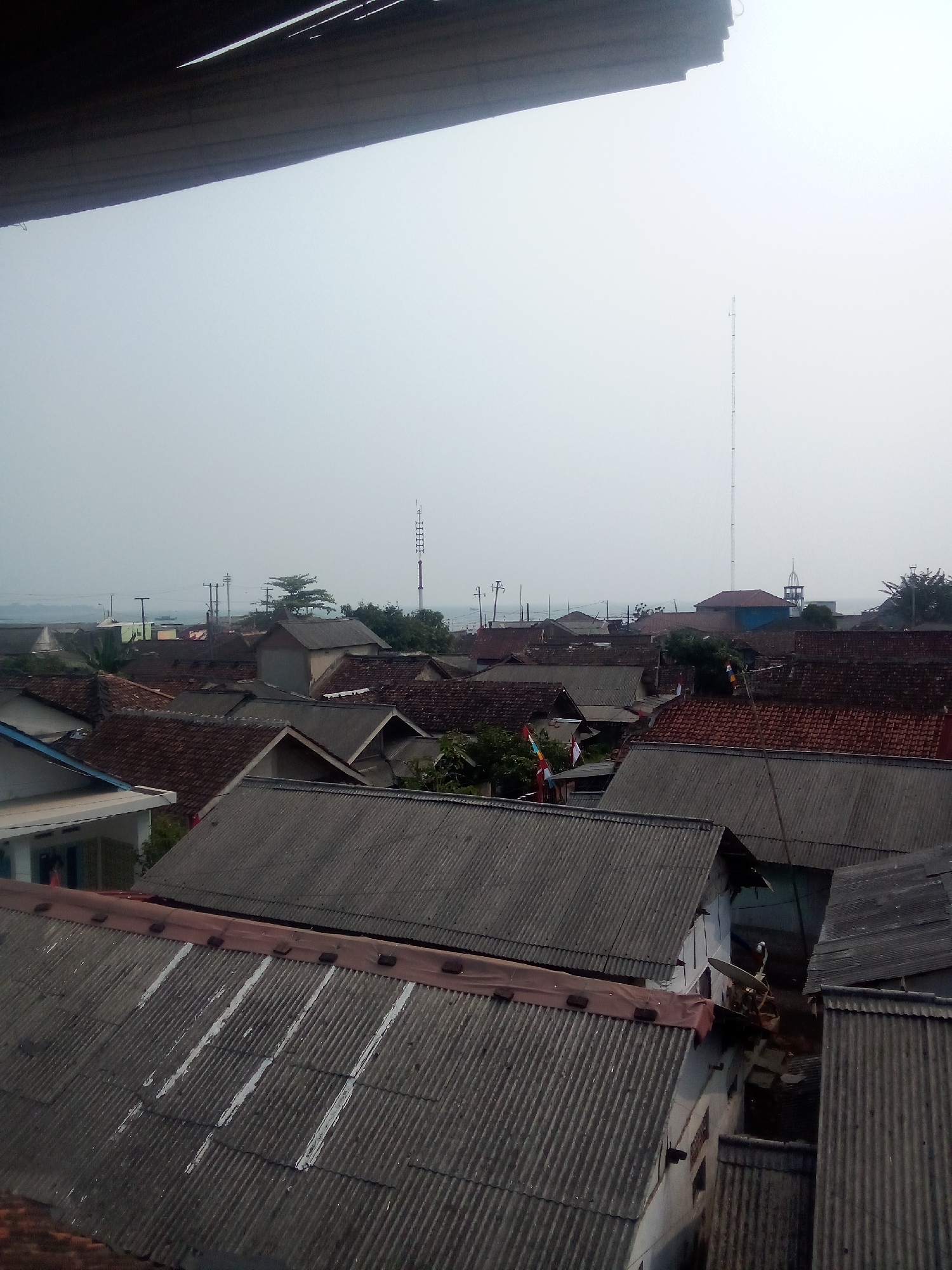 The image shows an aerial view of a residential area with many houses. The roofs are predominantly made of corrugated metal and red tiles. In the background, there is a body of water, possibly the sea or a large river, with a hazy sky. A tall communication tower is visible in the distance, along with some smaller structures and trees. There are also some power lines and poles scattered throughout the scene. The foreground features several corrugated metal roofs, some of which appear to be aged and possibly in need of repair, with white paint markings on some of them. The overall impression is that of a densely populated coastal or riverside community. The location is identified as Teluk, Indonesia. There are no visible people or animals in the image.